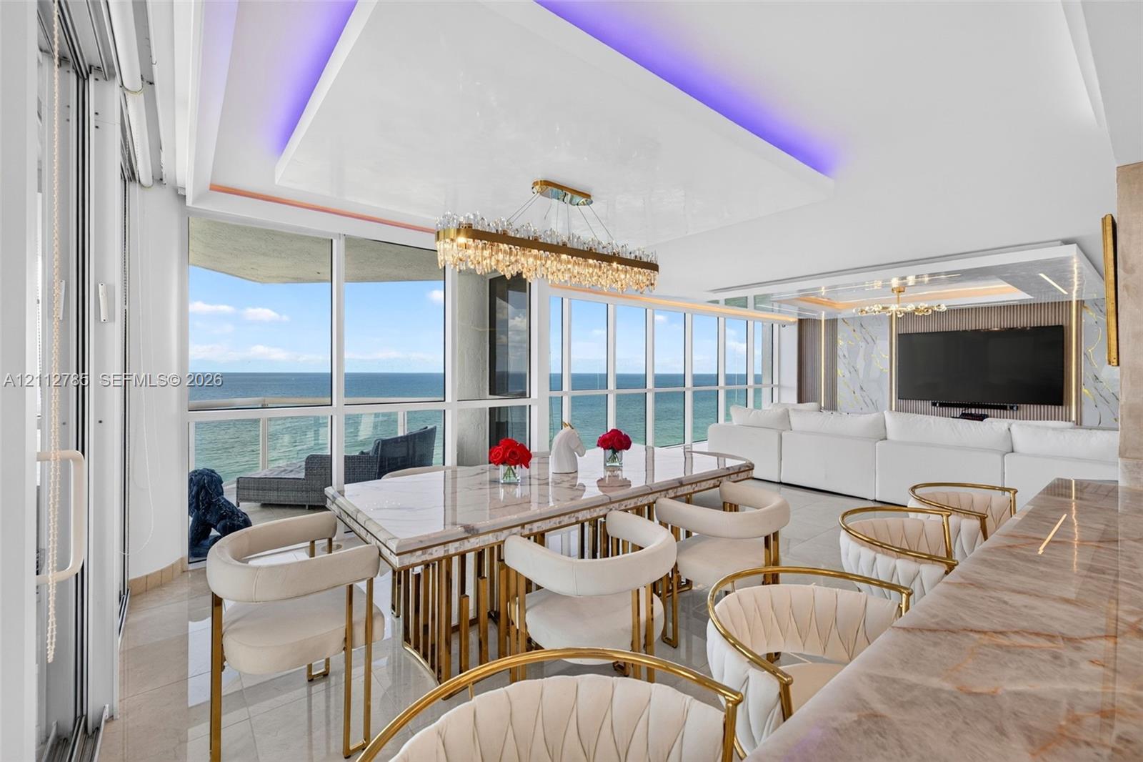 Floor-to-ceiling windows with coastal horizon, recessed tray ceiling with integrated perimeter lighting, multi-tiered crystal linear chandelier, polished stone-finish surfaces, and large-format porcelain tile flooring
