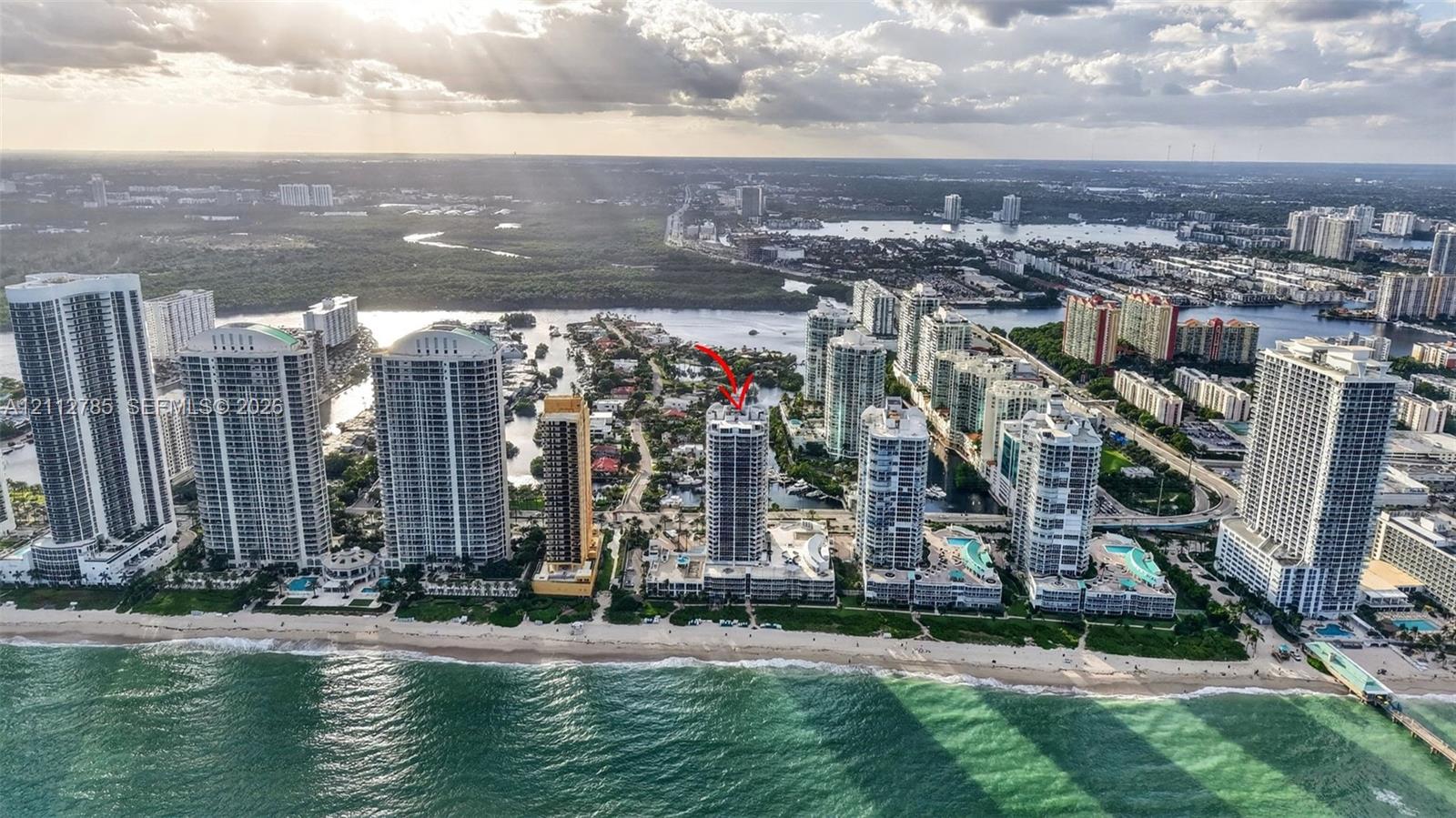 Aerial perspective of a high-rise tower positioned along a coastal shoreline, featuring direct ocean access, integrated building amenities, proximity to local waterways, and surrounding urban infrastructure