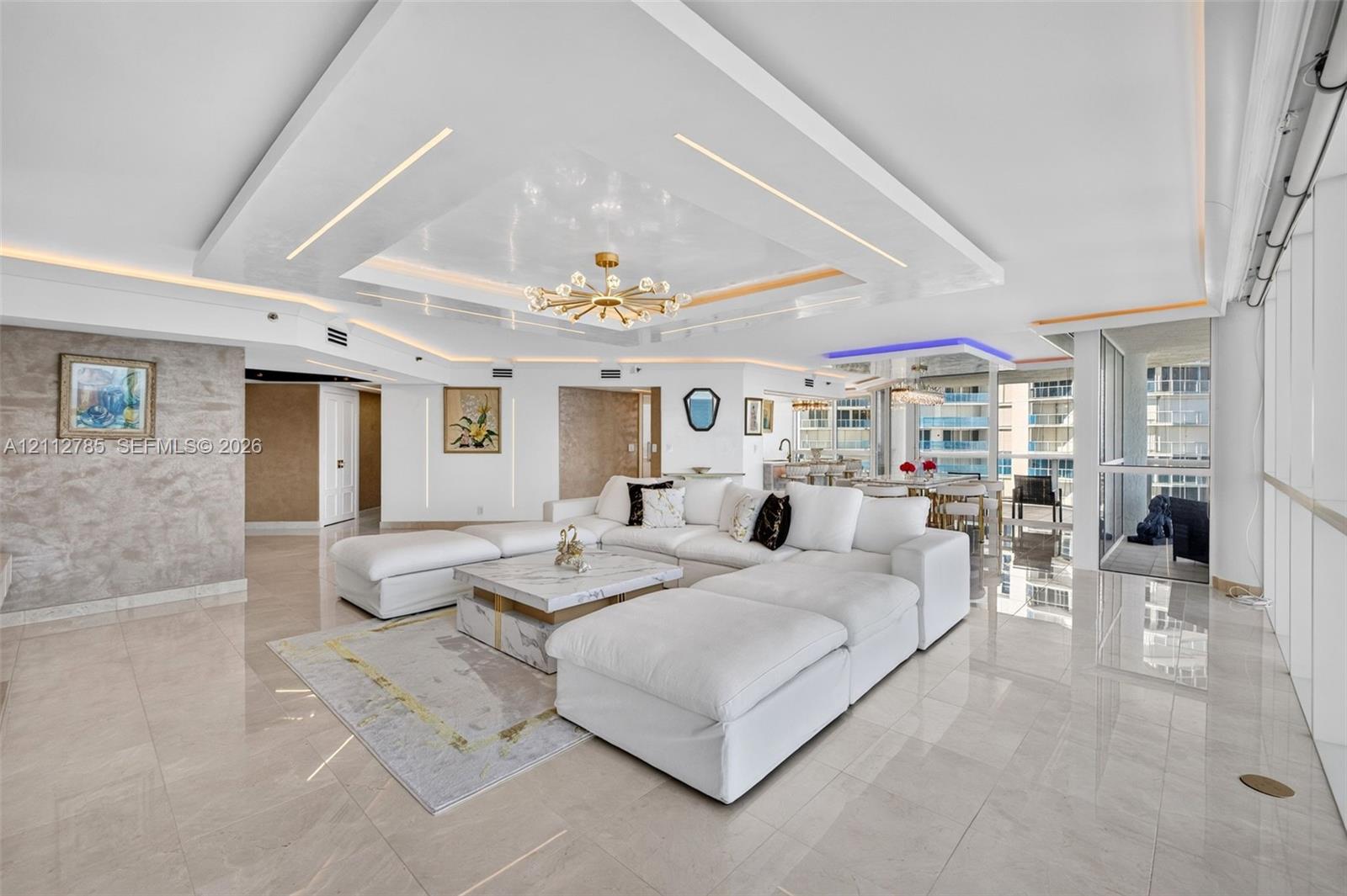 Open-concept living area featuring a custom recessed tray ceiling with integrated lighting, polished stone-finish flooring, floor-to-ceiling windows, and a textural wall finish