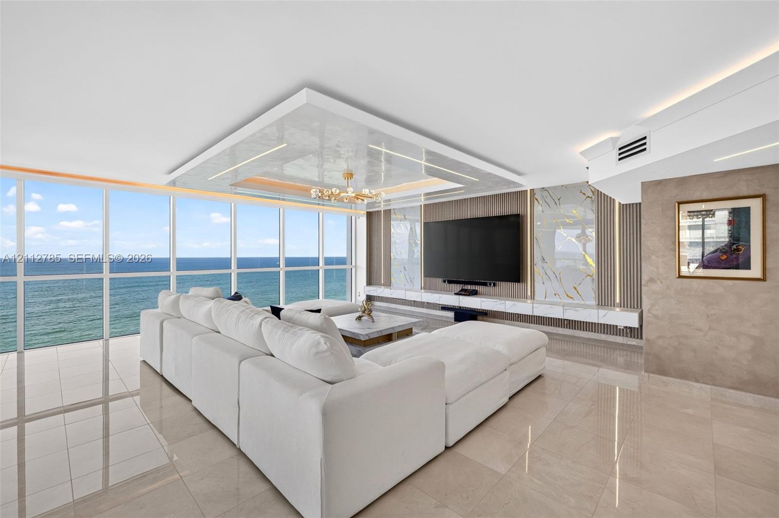 Floor-to-ceiling windows featuring ocean scenery, expansive polished tile flooring, tray ceiling with recessed lighting, and a built-in media wall with stone-finish paneling and under-cabinet illumination Floor-to-ceiling windows featuring ocean scenery, expansive polished tile flooring, tray ceiling with recessed lighting, and a built-in media wall with stone-finish paneling and under-cabinet illumination