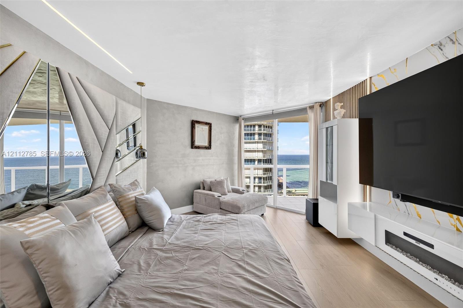 Wood-finish flooring, floor-to-ceiling windows with coastal scenery, accent wall with decorative mirror panels, wall-mounted linear fireplace, and integrated wood-slat wall paneling