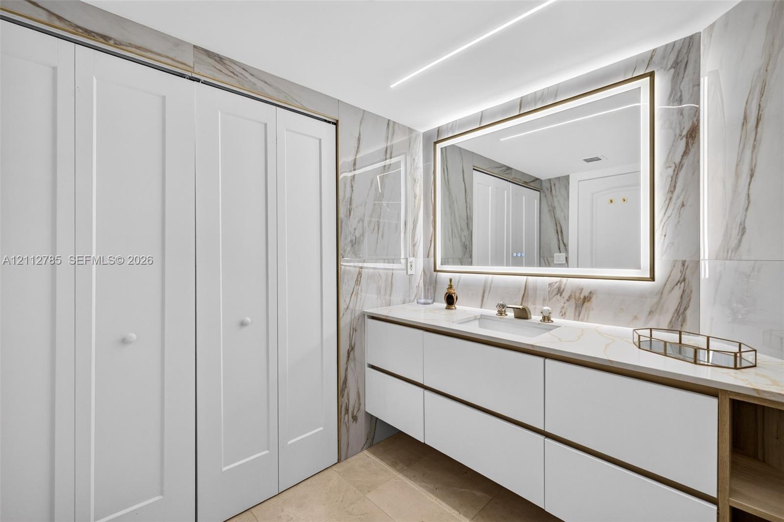 Floating vanity featuring flat-panel drawers, light-toned stone-finish countertop, integrated under-mount sink, LED-backlit mirror, and floor-to-ceiling porcelain tile wall cladding