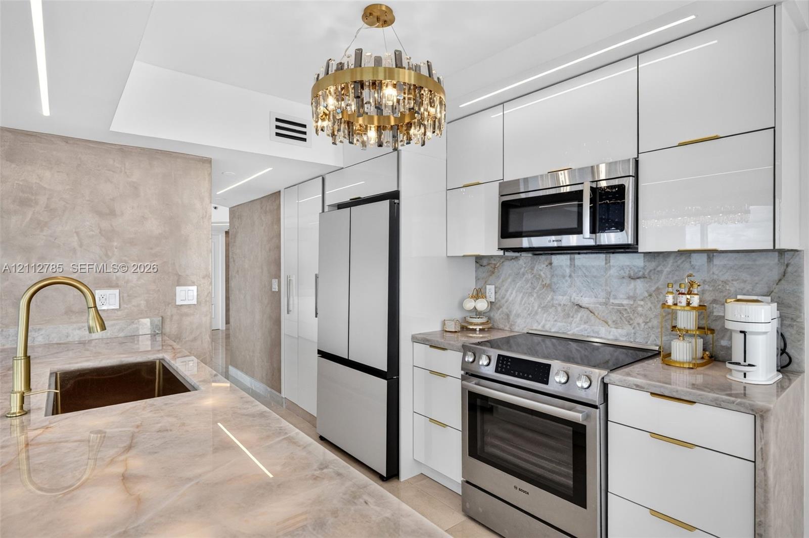 Glossy white cabinetry with gold-tone hardware, stone-finish countertops and backsplash, professional-grade stainless steel appliances, and a circular crystal chandelier