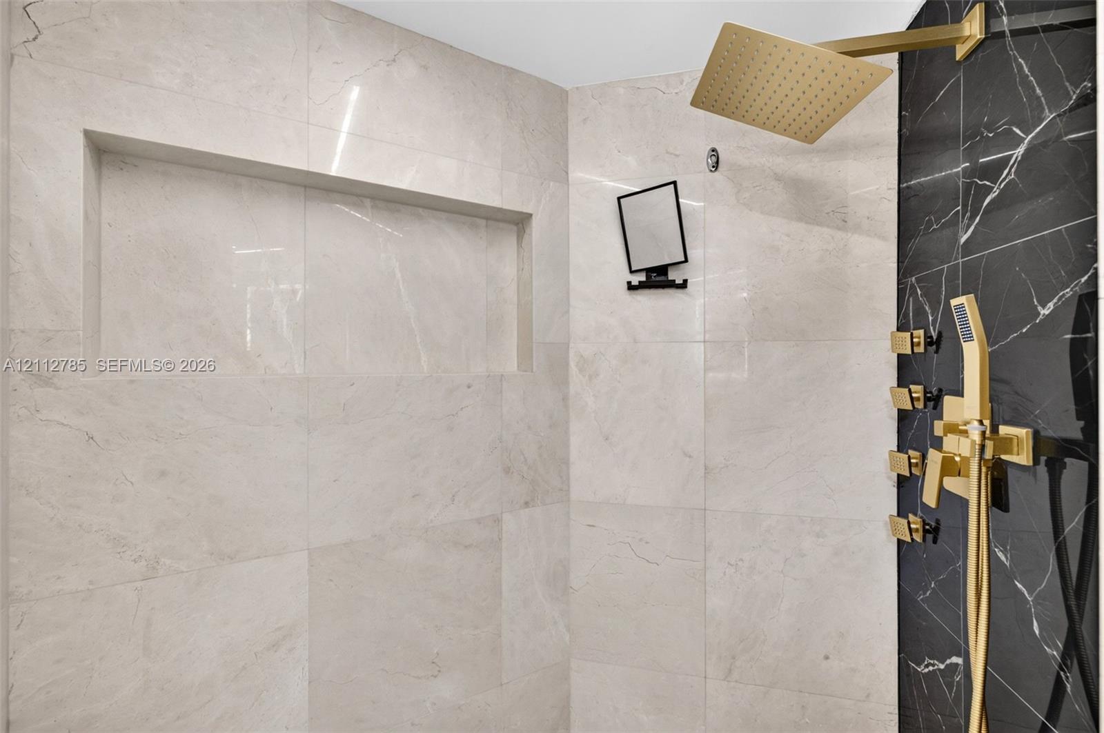 Walk-in shower featuring large-format light-tone tile, a contrasting black tile accent wall, brass finish rainfall shower head, integrated wall niche, and matching brass multi-function hand-held sprayer