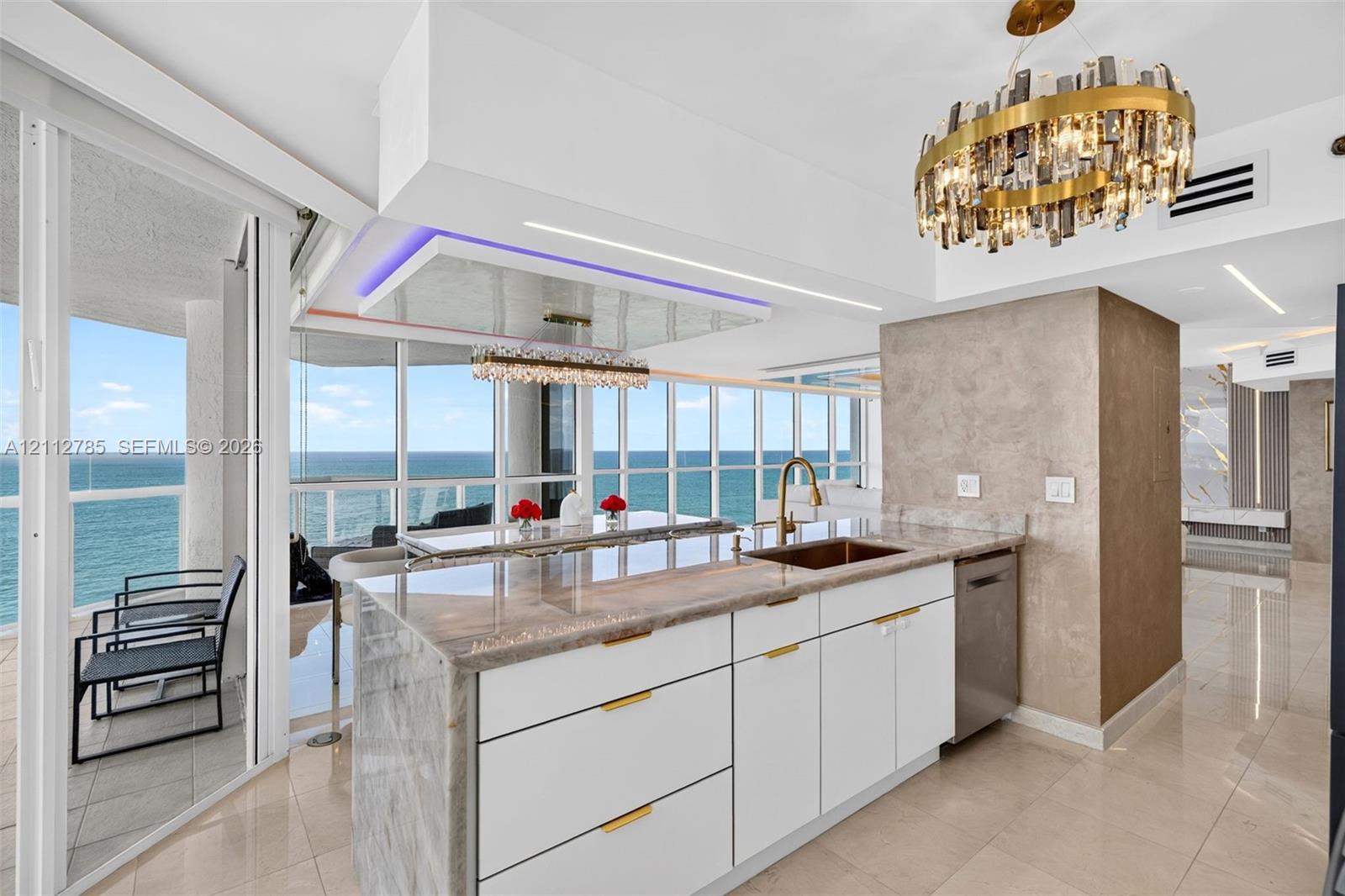 Kitchen island featuring a stone waterfall countertop, integrated stainless steel sink, gold-tone hardware, white cabinetry, and polished tile flooring