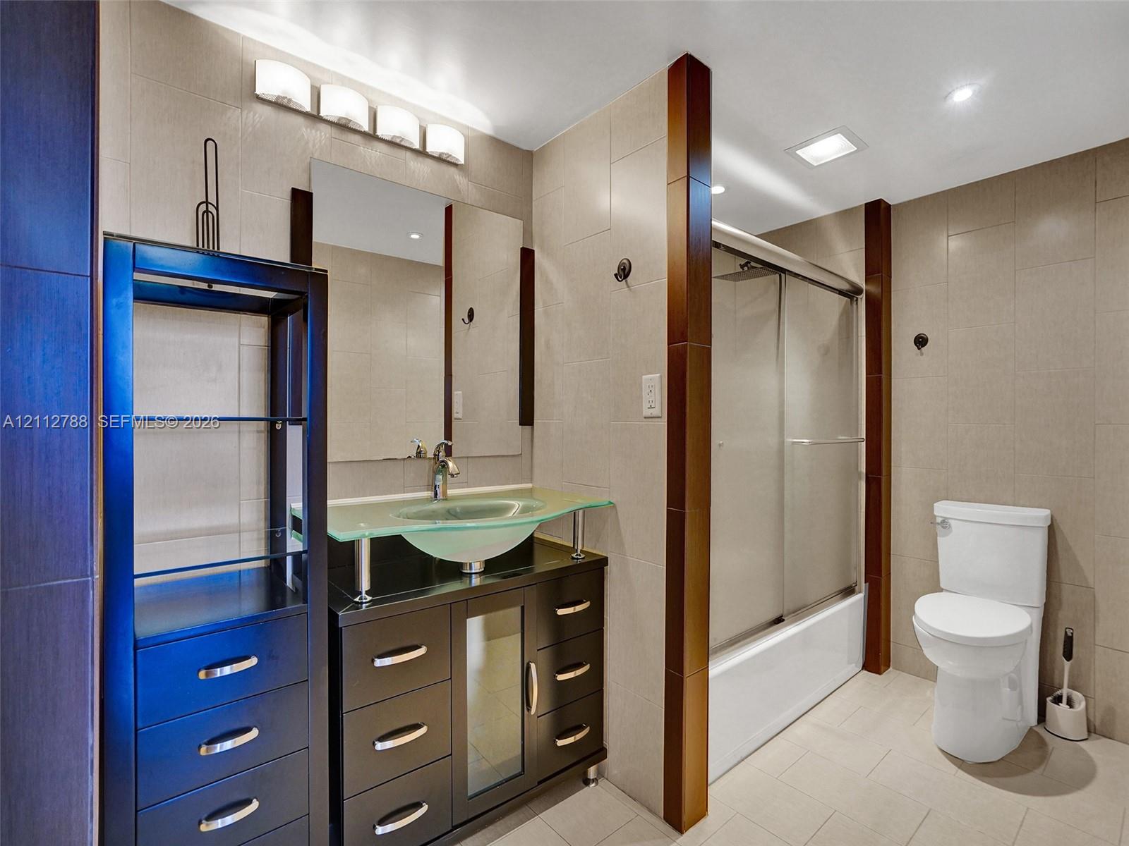Bathroom featuring a dark wood-finish vanity with a glass vessel sink, glass sliding shower door with tile surround, and neutral tile flooring