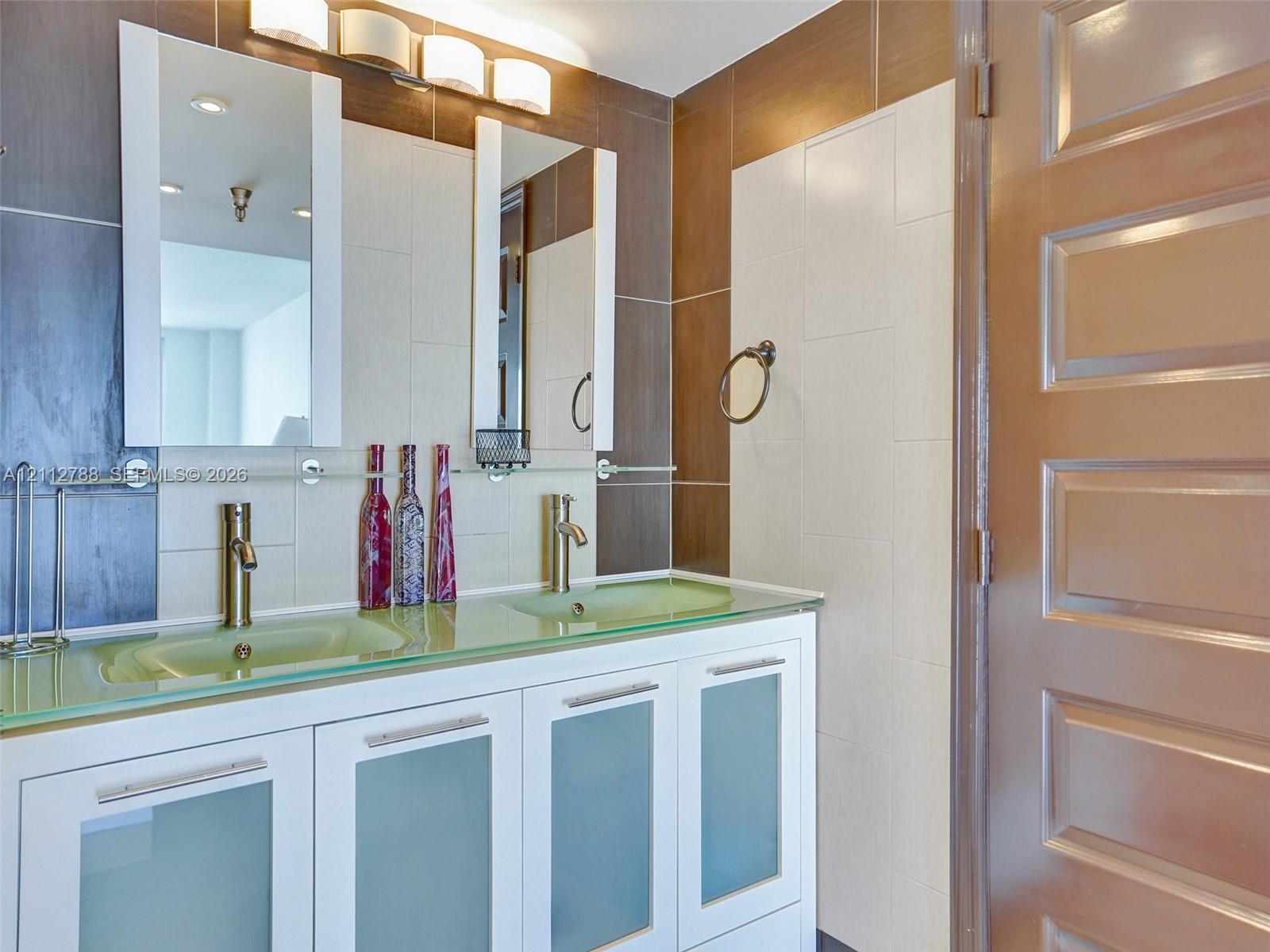 Double vanity with glass countertop, integrated sinks, white cabinetry with frosted glass inserts, metallic hardware, and dual vertical wall-mounted mirrors