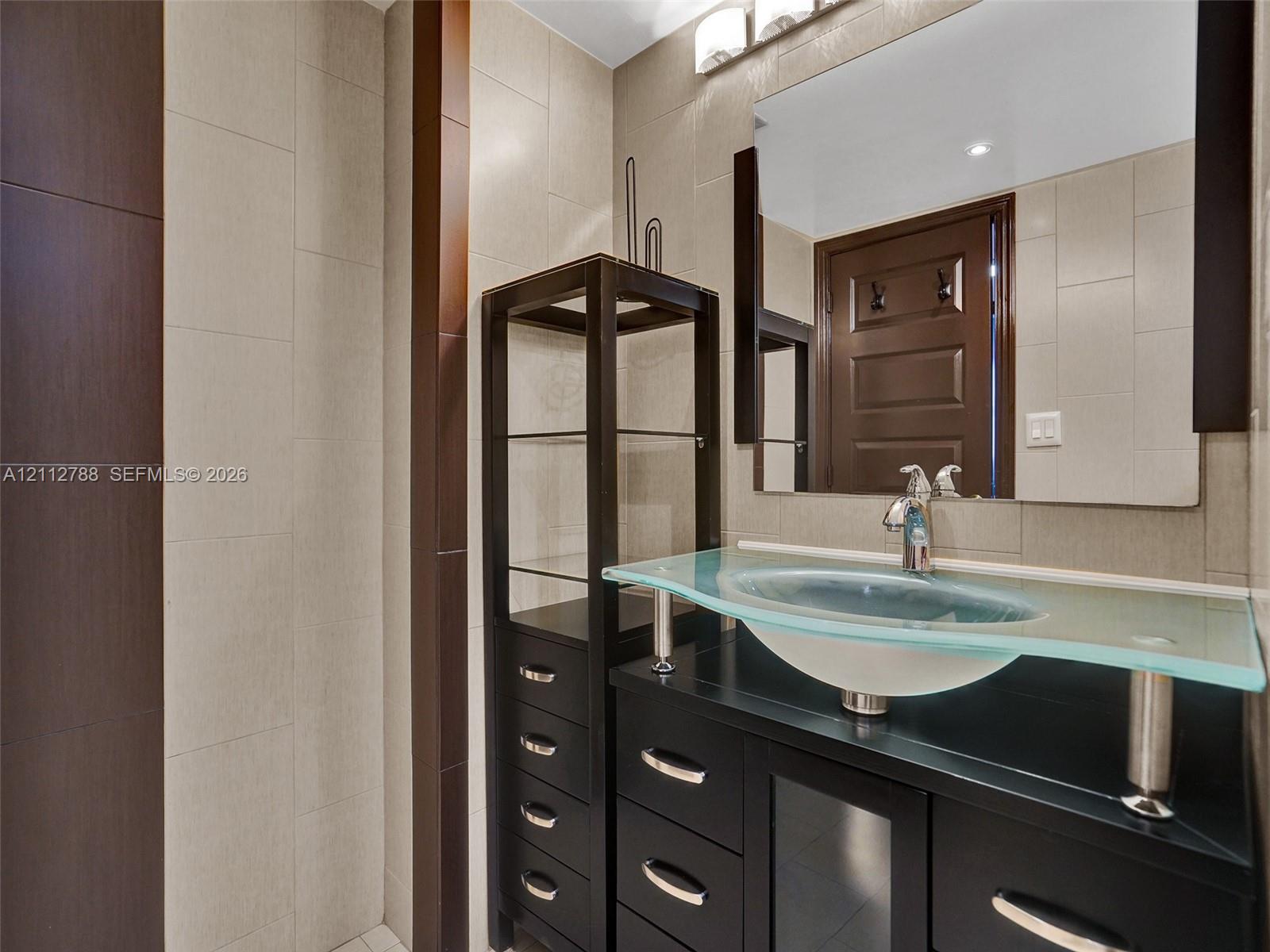 Modern vanity featuring a glass countertop, integrated vessel sink, polished chrome faucet, dark cabinetry, and floor-to-ceiling tile work