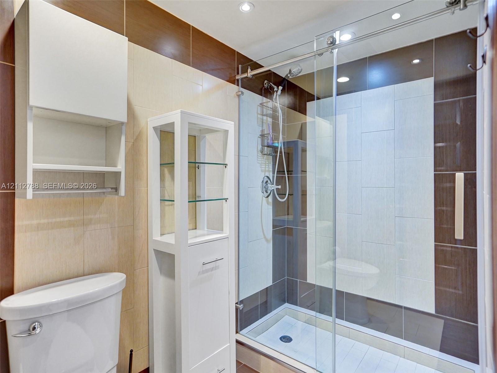Walk-in shower featuring a sliding glass enclosure, integrated wall shelving, and dual-tone ceramic tile surround with built-in white cabinetry and recessed lighting