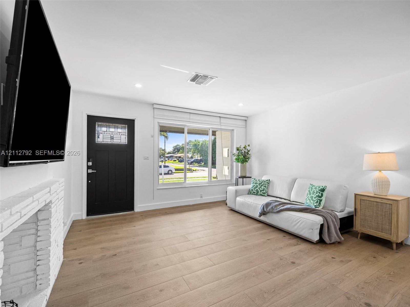 Living space featuring wood-finish flooring, a painted brick fireplace, a black front door with an integrated window, and a large picture window