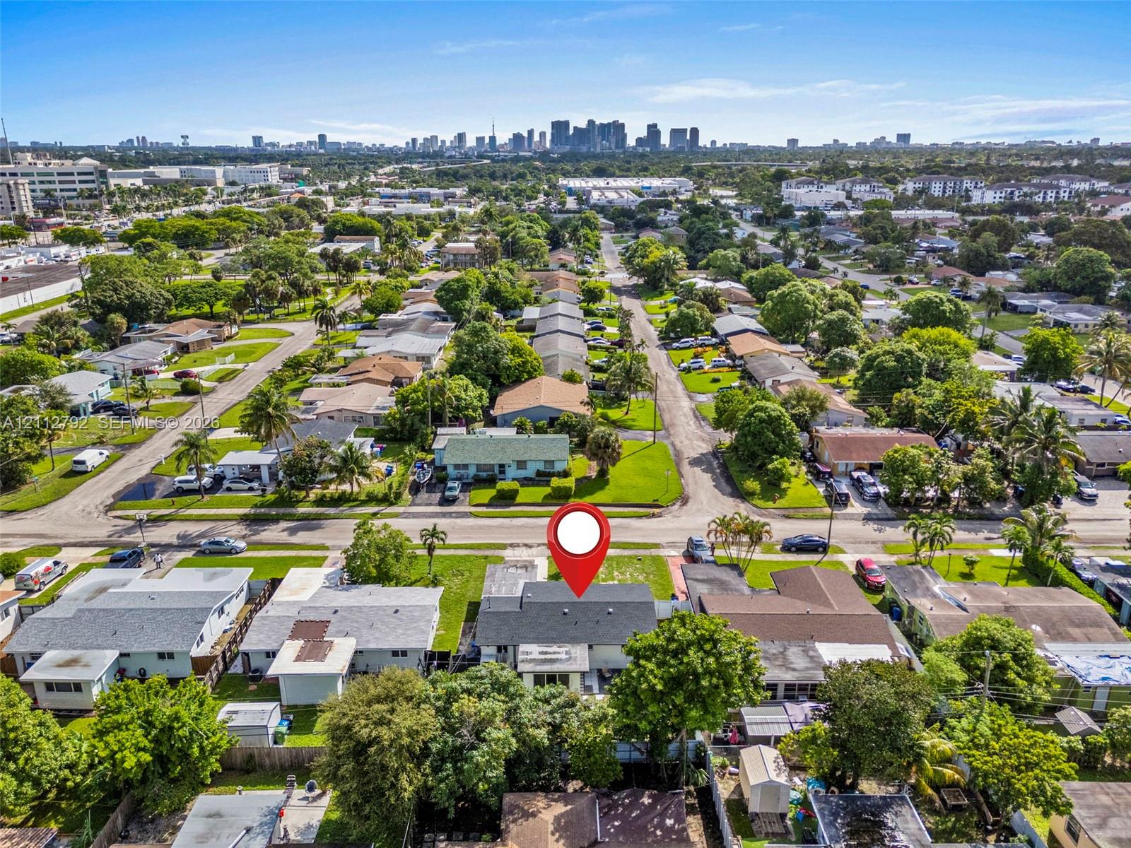 Aerial perspective showcasing a residential property with a shingle roof and mature landscaping, including palm trees, situated in an established neighborhood with a distant cityscape backdrop