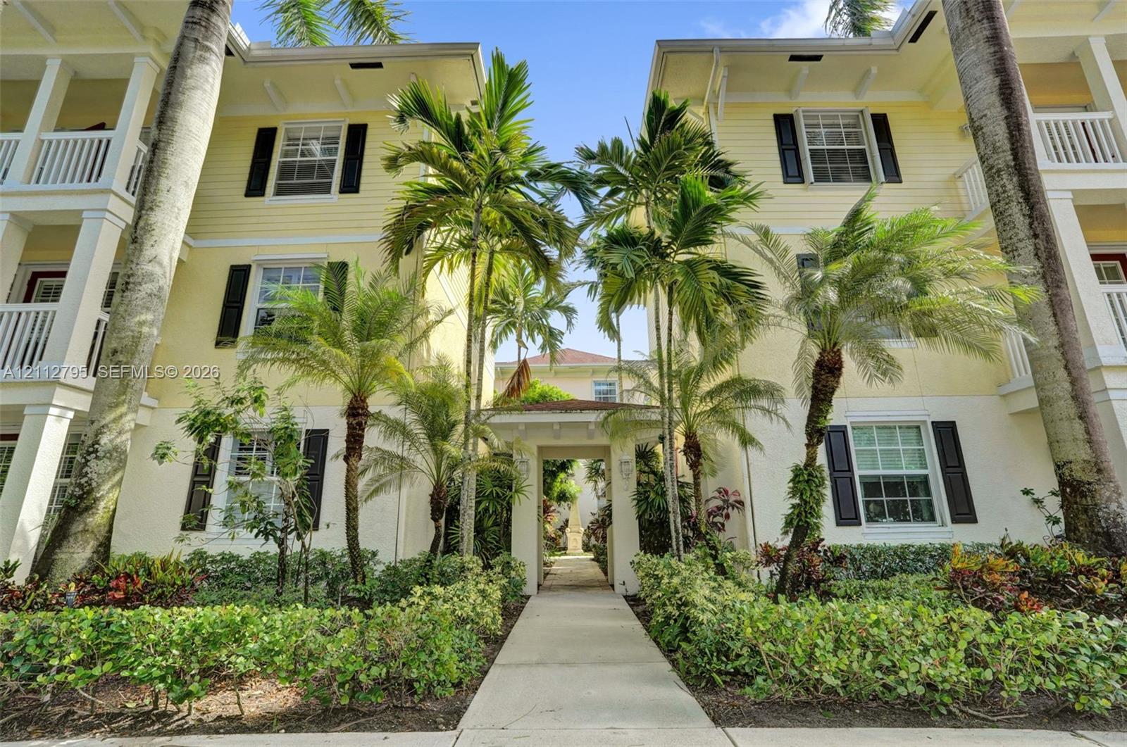 Multi-story exterior featuring light-colored horizontal siding, dark decorative shutters, covered balconies with white railing, and mature palm landscaping