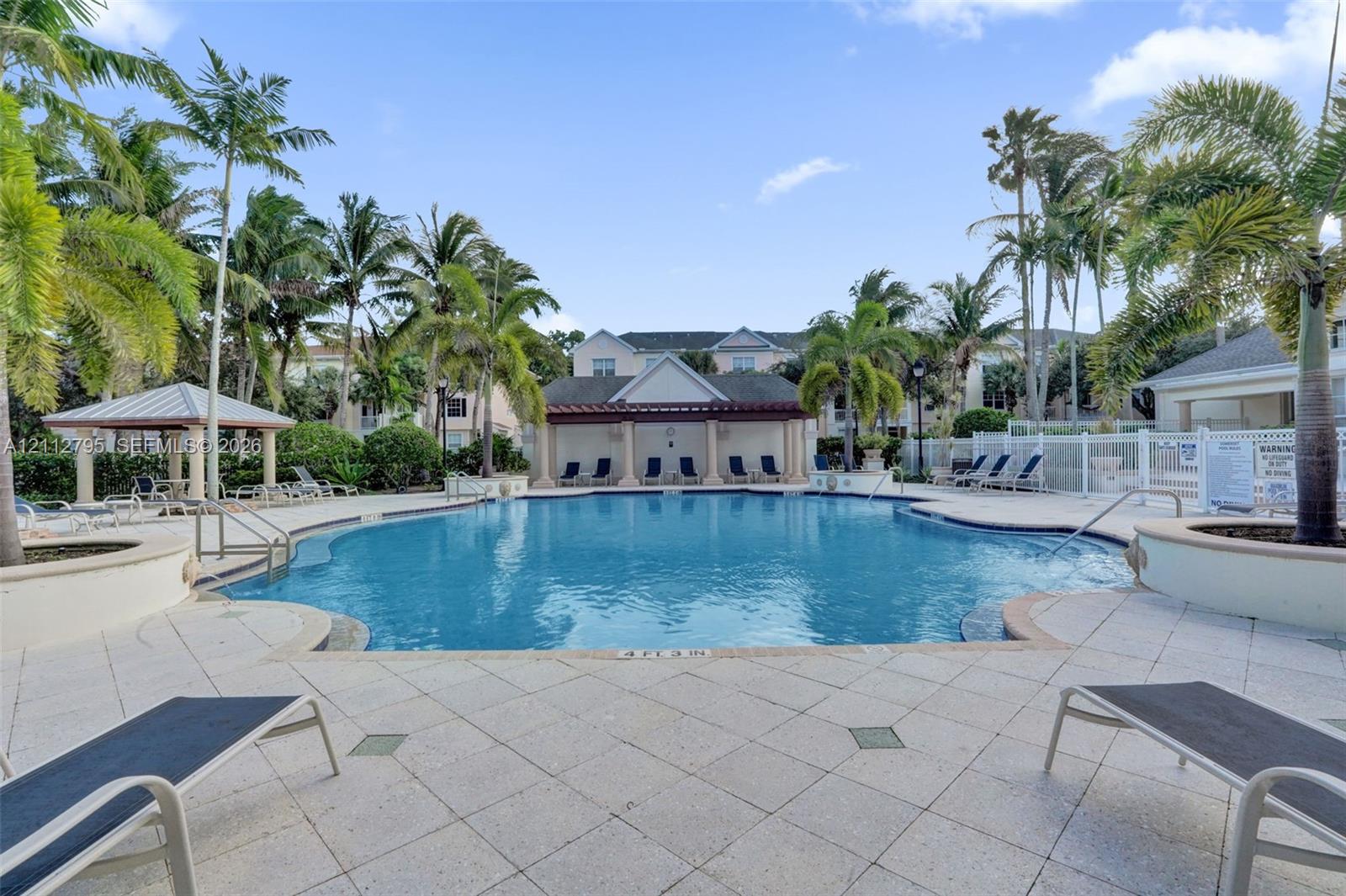 Large community swimming pool featuring a stone-paver deck, multiple integrated spas, a covered cabana with columns, and mature palm trees