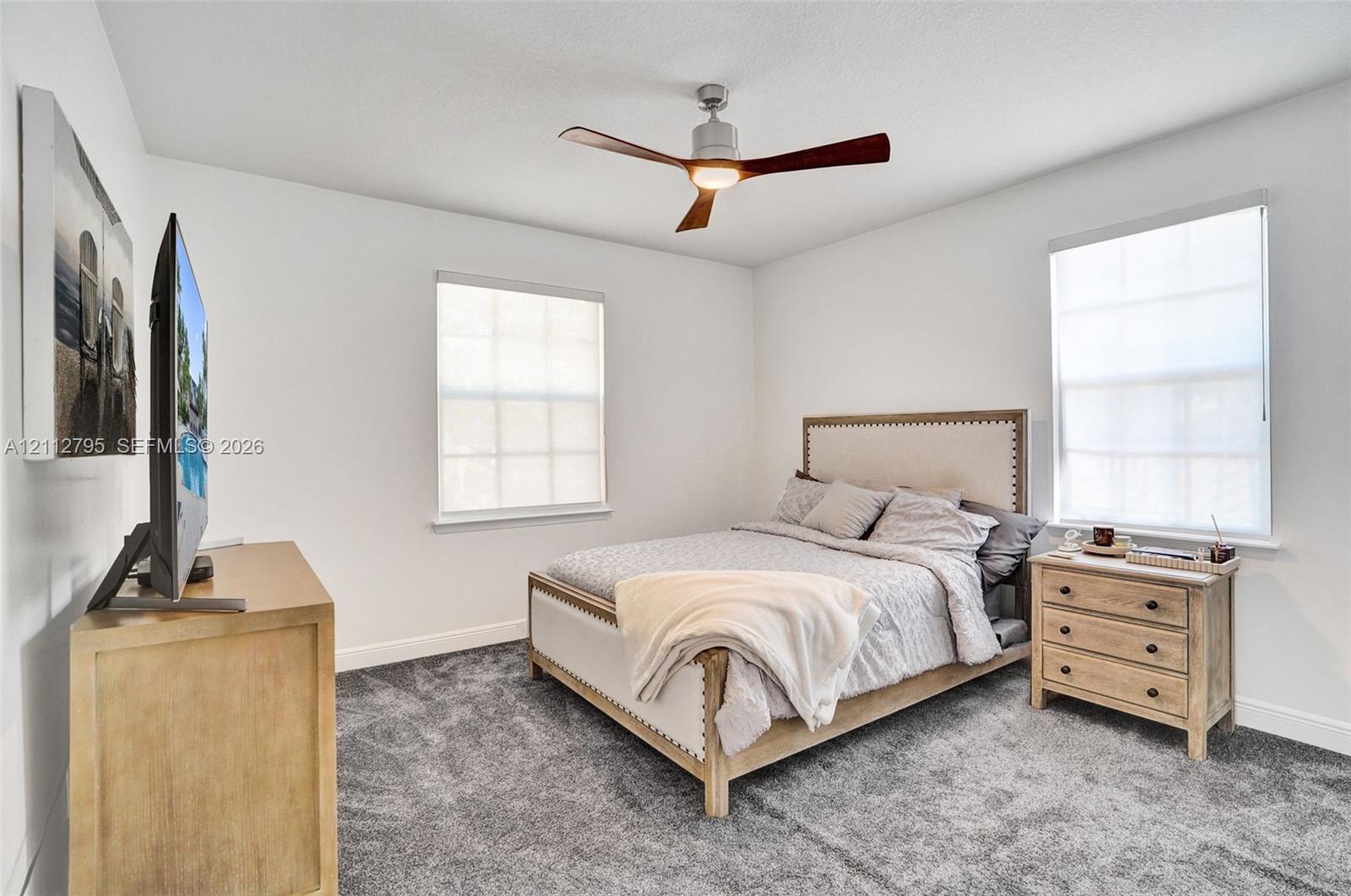 Neutral-toned room featuring grey wall-to-wall carpeting, dual windows with roller shades, and a ceiling fan with integrated lighting