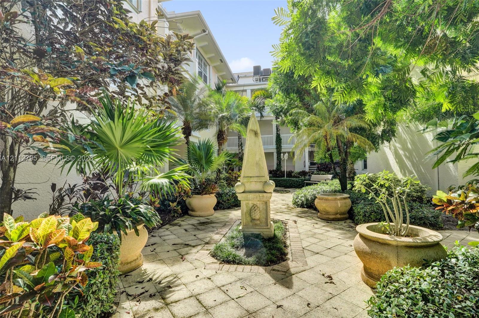 Courtyard featuring stone paver flooring, central stone obelisk monument, mature trees, ornamental palms, and oversized stone planters