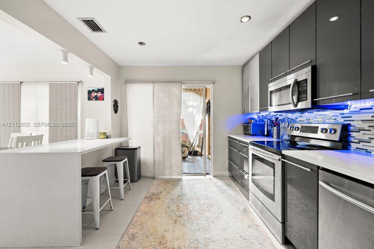 Kitchen featuring high-gloss charcoal cabinetry, stainless steel appliances, quartz countertops, a stone-tile backsplash, and a breakfast bar