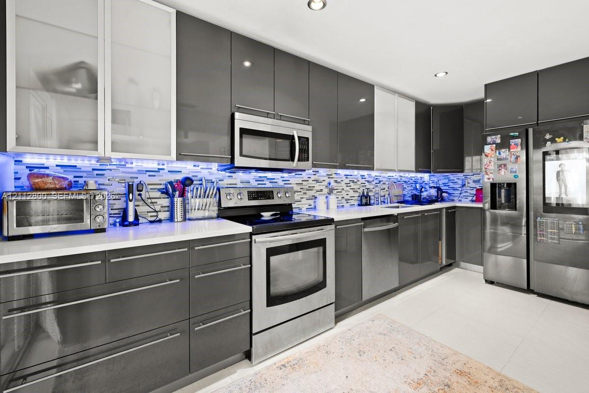 Kitchen featuring high-gloss charcoal cabinetry, glass-front upper cabinets, stainless steel appliances, a multi-tone glass tile backsplash, and quartz countertops