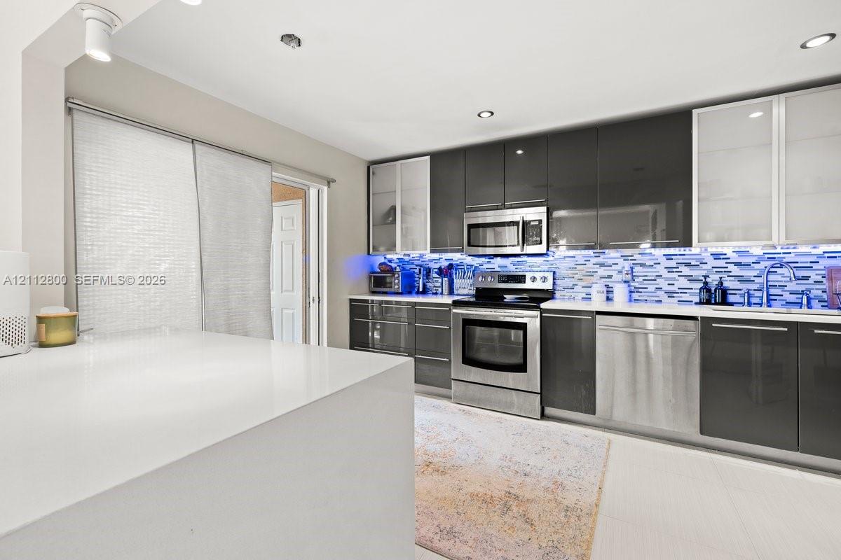 Modern kitchen featuring high-gloss charcoal cabinetry, stainless steel appliances, a white quartz waterfall-edge island, glass-front upper cabinets, and a multi-toned mosaic tile backsplash