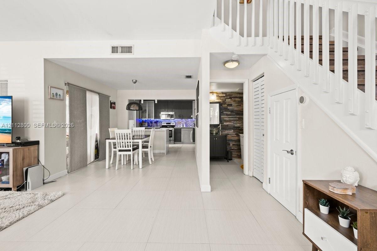 Large-format ceramic tile flooring, staircase with white railings, kitchen featuring dark cabinetry and stainless-steel appliances, and stone-accented wall in the secondary room