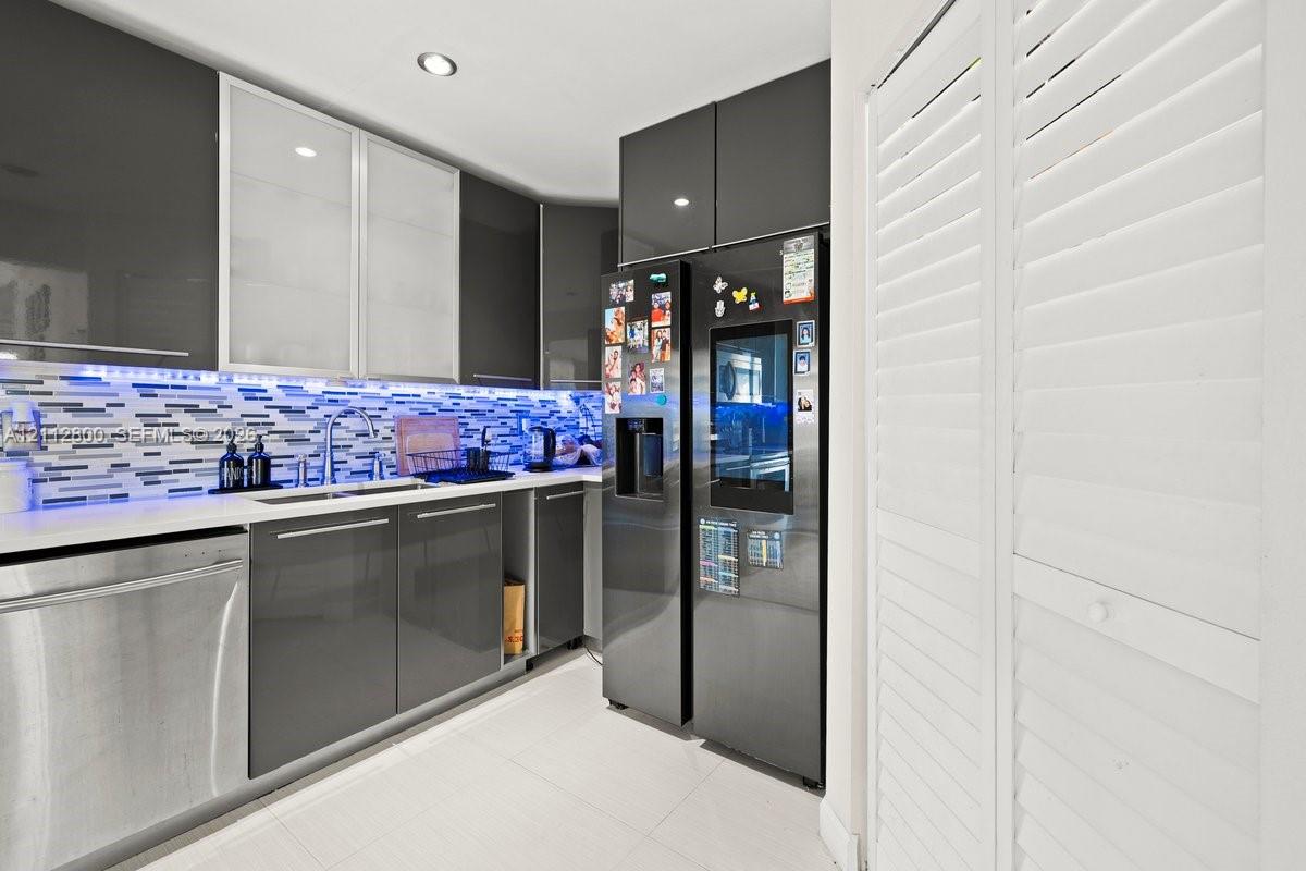 Kitchen featuring high-gloss charcoal cabinetry, glass-fronted upper cabinets, multi-toned horizontal tile backsplash, stainless steel dishwasher, and floor-to-ceiling white louvered closet doors