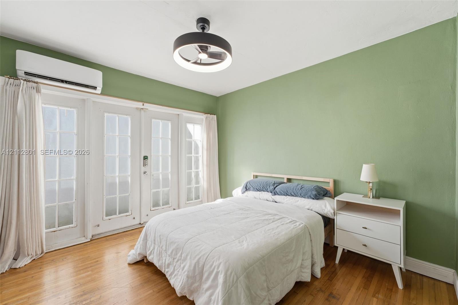 Wood-finish flooring, wall-mounted ductless climate control unit, double French doors with glass panes, and a modern drum-style ceiling fan