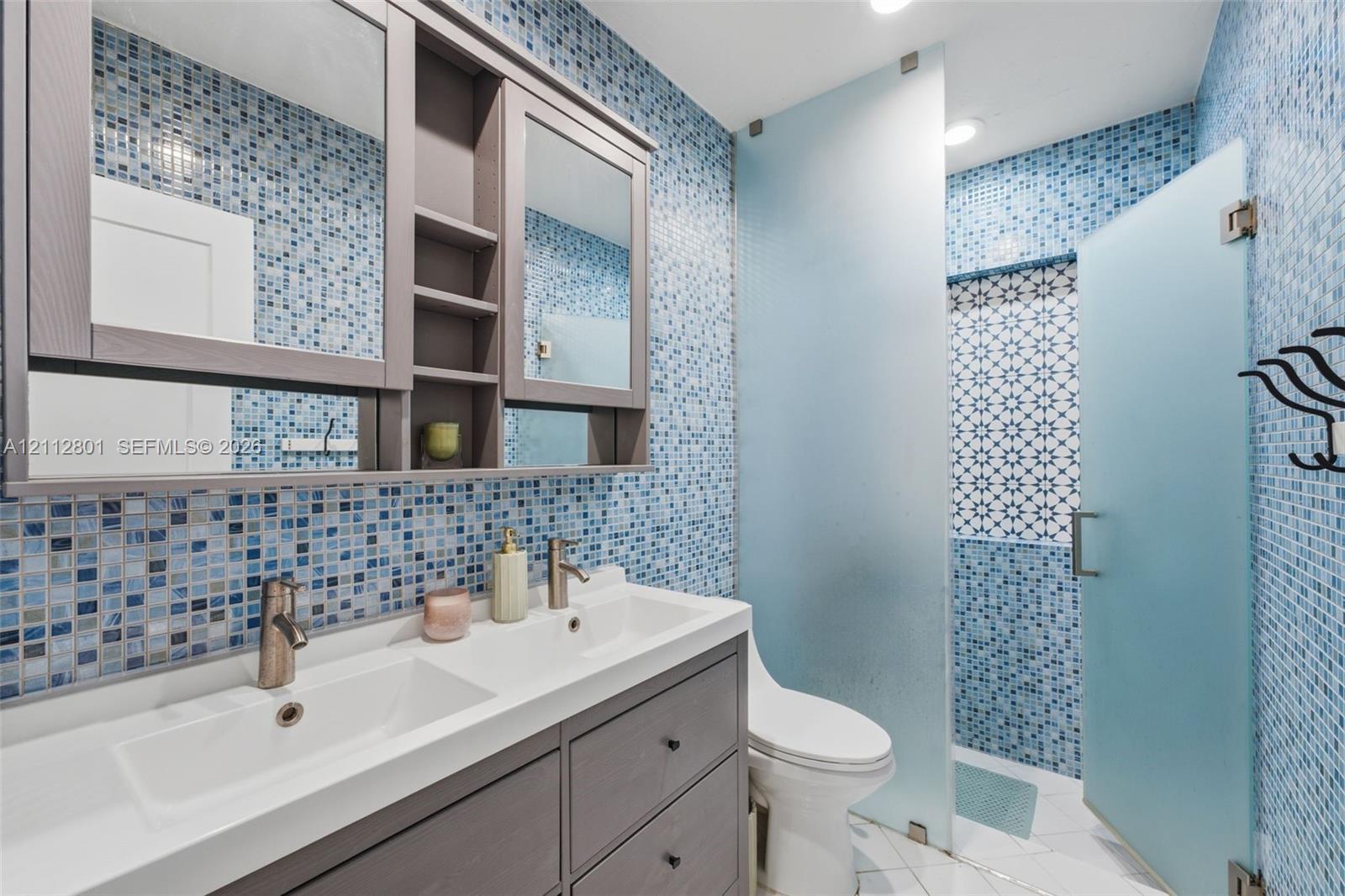 Double vanity featuring a white integrated countertop with dual basins and matte-finish hardware, built-in medicine cabinets, full-height blue glass mosaic tile walls, and a walk-in shower enclosure with frosted glass panels
