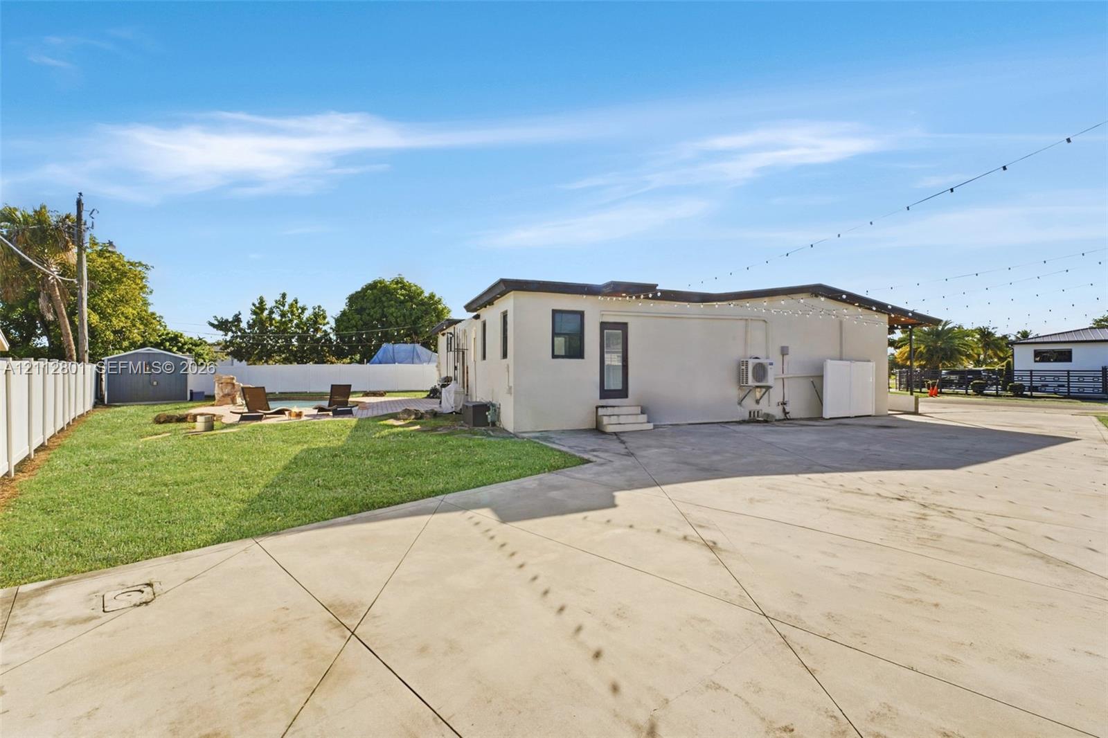 Expansive concrete hardscaping, manicured lawn, white vinyl fencing, storage shed, and exterior string lighting