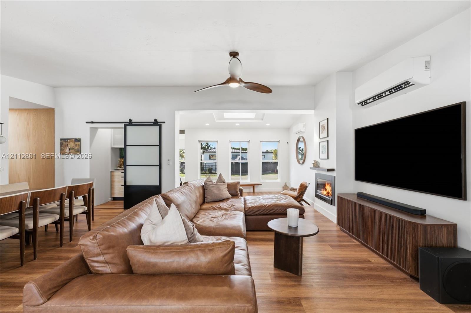 Open-concept interior featuring wood-finish flooring, a contemporary ceiling fan, a wall-mounted fireplace, a black-framed sliding barn door with frosted panels, and a wall-mounted mini-split HVAC unit