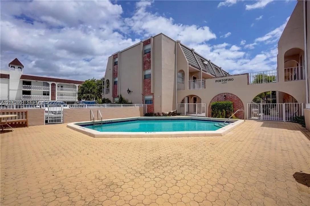 Exterior common area featuring an inground swimming pool with metal ladder access, surrounded by a paved deck and enclosed by a white security fence Exterior common area featuring an inground swimming pool with metal ladder access, surrounded by a paved deck and enclosed by a white security fence
