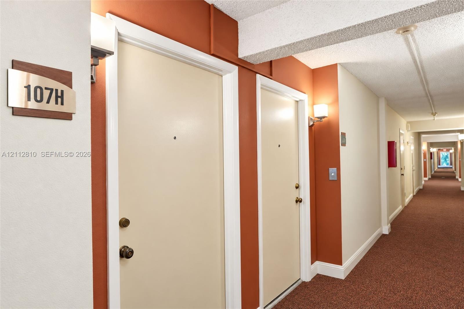 Interior hallway featuring wall-mounted light fixtures, carpeted flooring, and neutral-toned doors with metallic hardware Interior hallway featuring wall-mounted light fixtures, carpeted flooring, and neutral-toned doors with metallic hardware