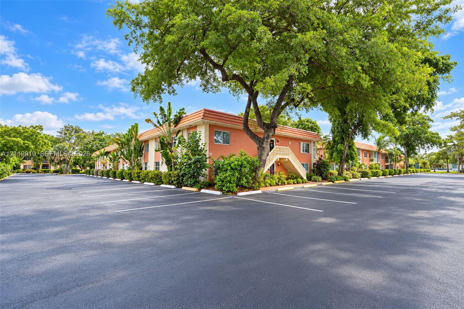 Multi-story building featuring stucco exterior walls, a tile roof, paved parking area, and mature landscaping Multi-story building featuring stucco exterior walls, a tile roof, paved parking area, and mature landscaping