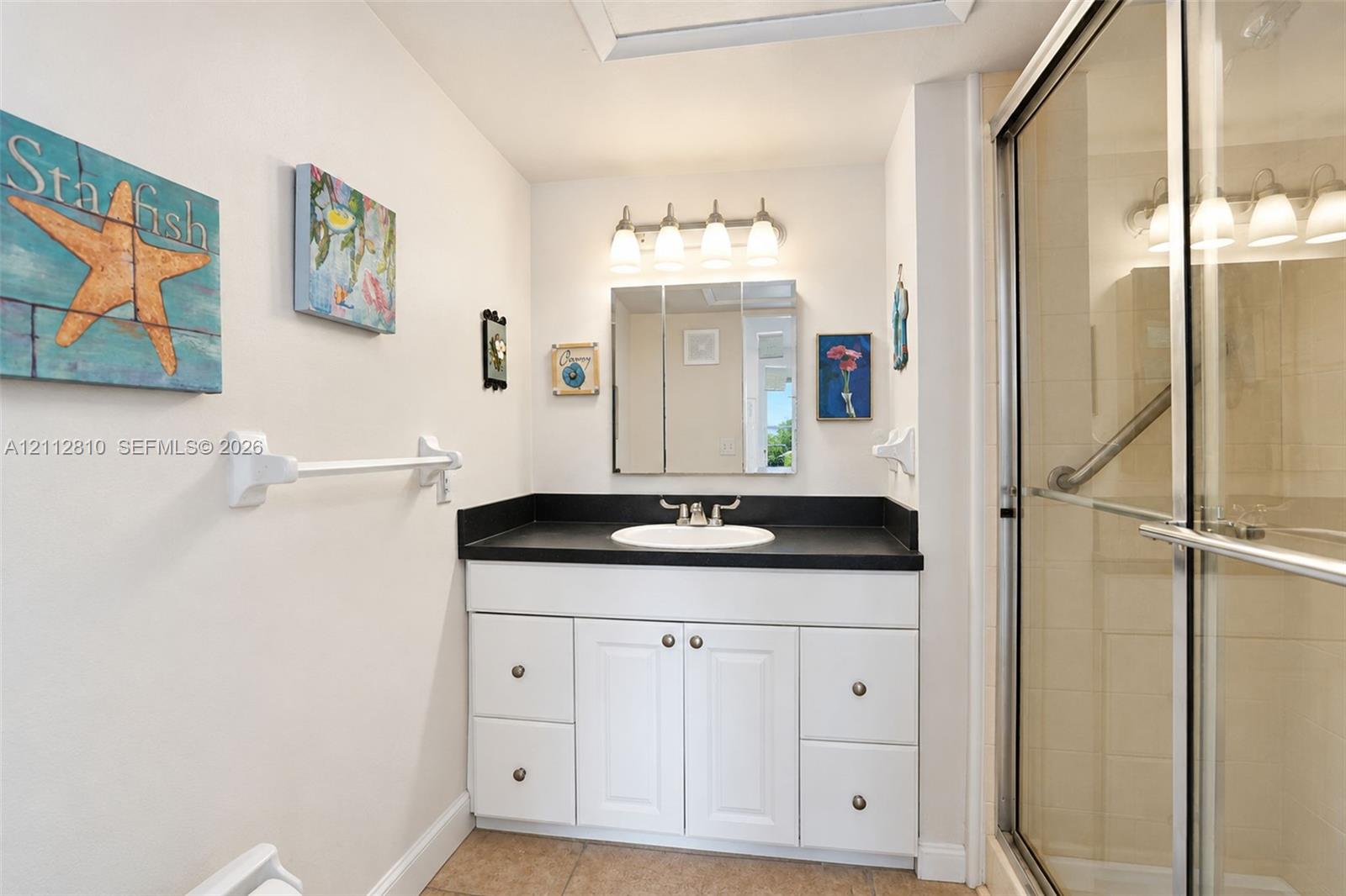 Bathroom vanity featuring white cabinetry, a dark countertop, an integrated sink, a triple-light fixture, and a tri-fold medicine cabinet, paired with a glass-enclosed shower