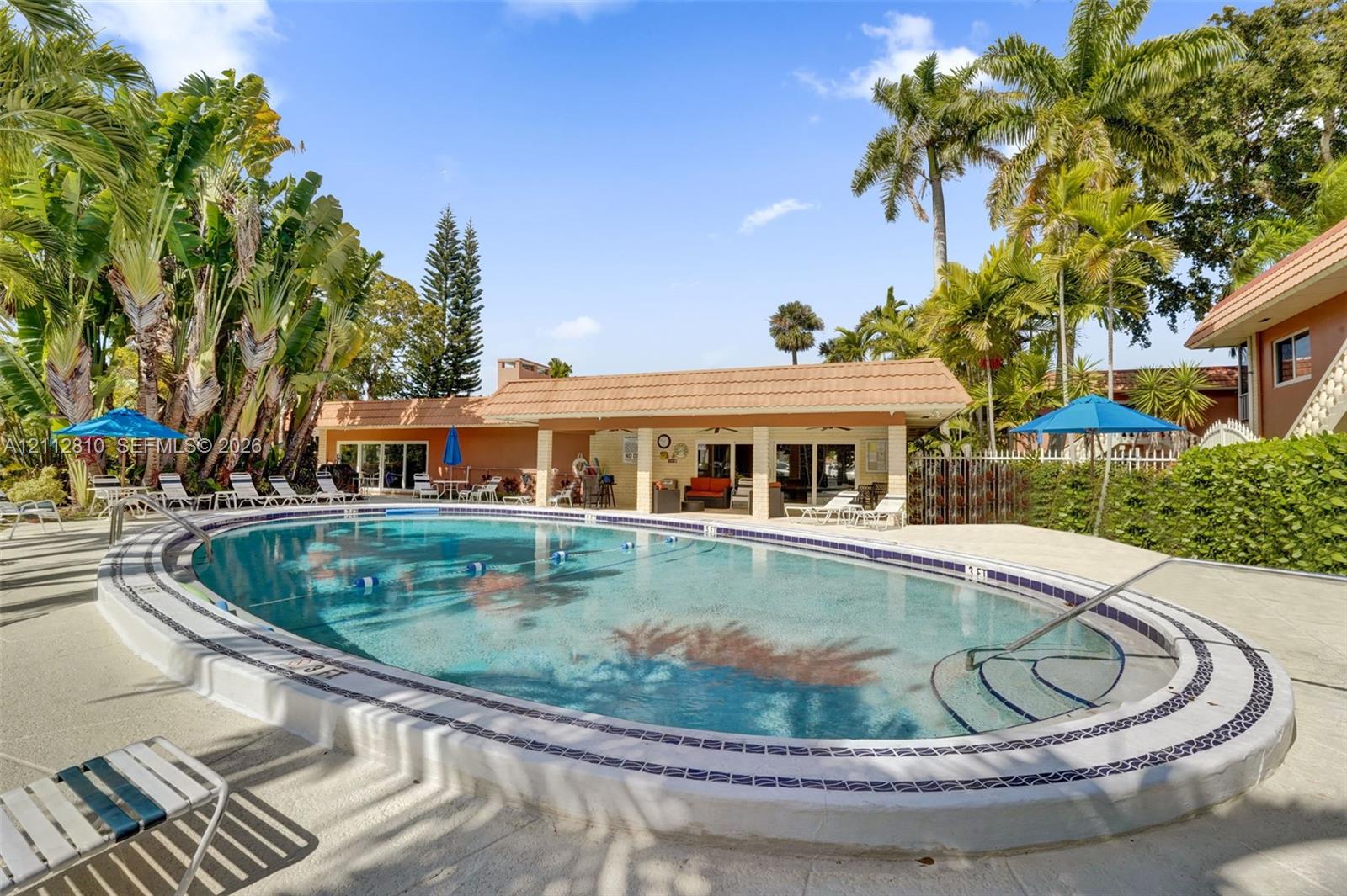 Oval swimming pool with tile border, recessed steps, and light-colored concrete decking, situated within a landscape featuring mature palm trees and a covered patio structure with tile roofing