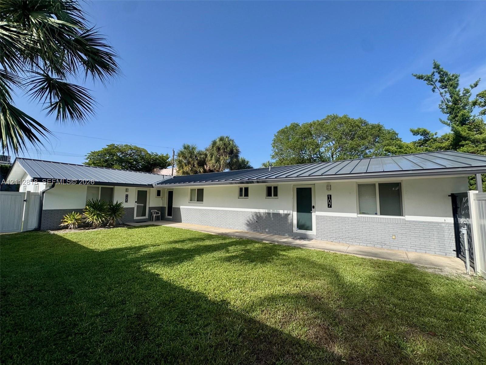 Single-story exterior featuring a metal roof, two-tone painted brick facade, manicured lawn, and perimeter privacy fencing Single-story exterior featuring a metal roof, two-tone painted brick facade, manicured lawn, and perimeter privacy fencing