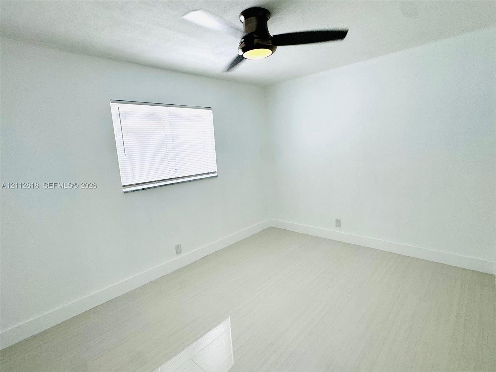 Interior room featuring light-toned wood-finish flooring, white walls, a dual-pane horizontal sliding window with blinds, and a matte black ceiling fan with an integrated light fixture