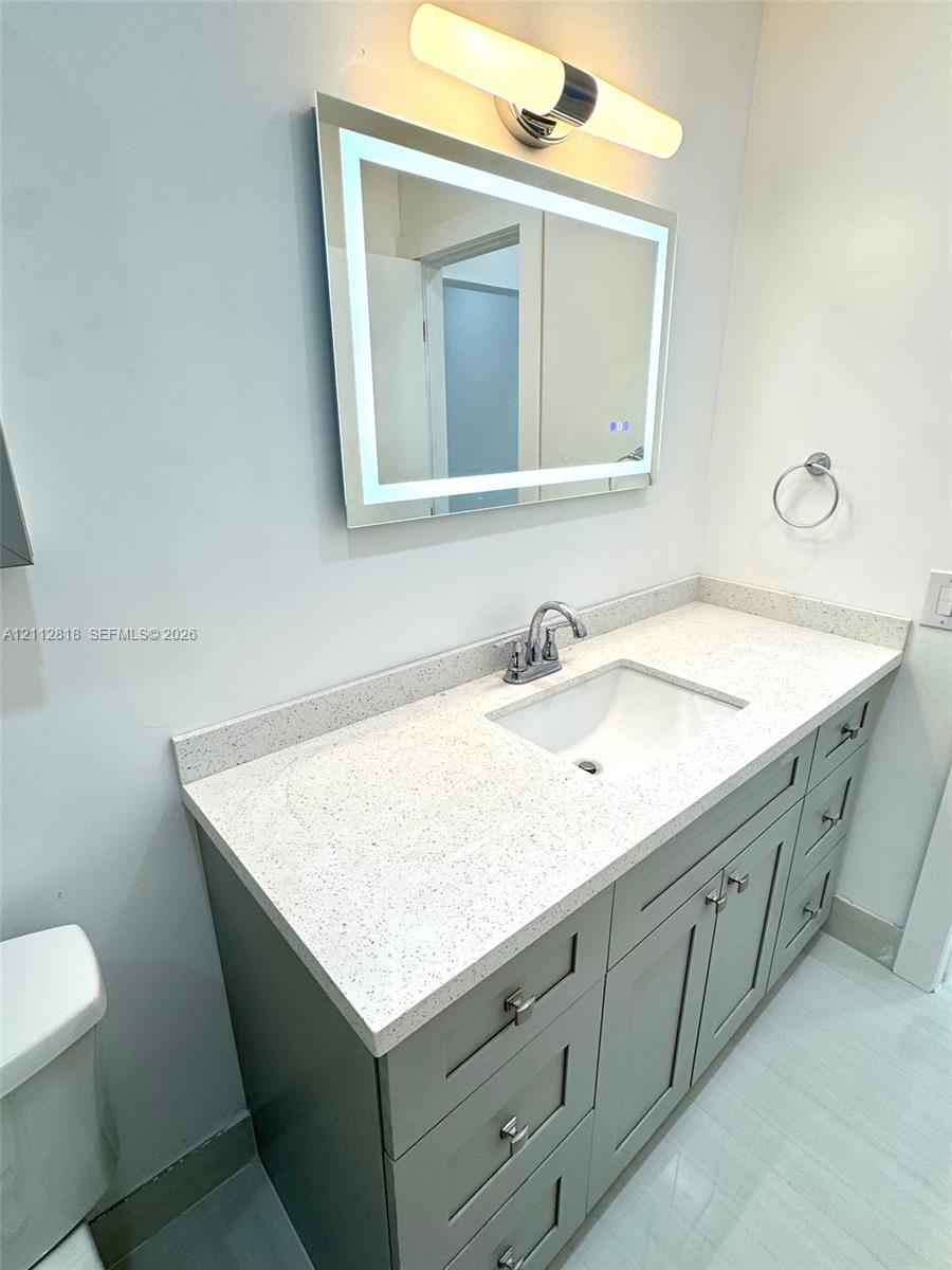Vanity featuring grey shaker-style cabinetry, a speckled composite countertop, an undermount sink, a rectangular LED-backlit mirror, and a contemporary wall-mounted light fixture