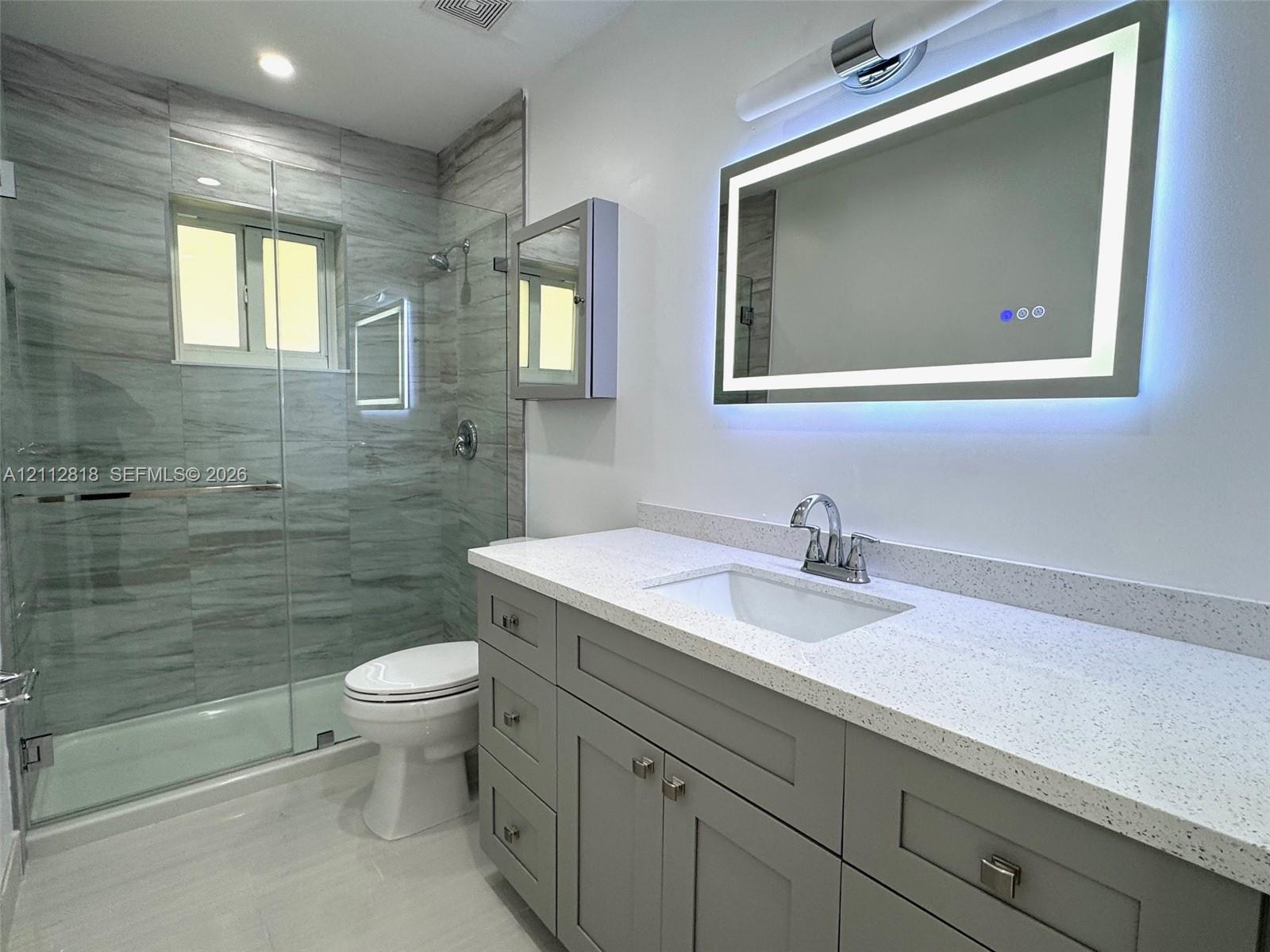 Shaker-style grey vanity with a speckled light countertop, illuminated rectangular mirror, and walk-in glass-enclosed shower featuring large-format tile surround