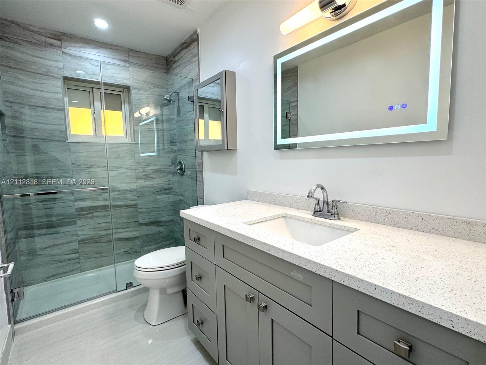Modern vanity with stone-composite countertop, undermount sink, and illuminated mirror featuring built-in cabinetry, walk-in shower with floor-to-ceiling tile, and frameless glass enclosure Modern vanity with stone-composite countertop, undermount sink, and illuminated mirror featuring built-in cabinetry, walk-in shower with floor-to-ceiling tile, and frameless glass enclosure