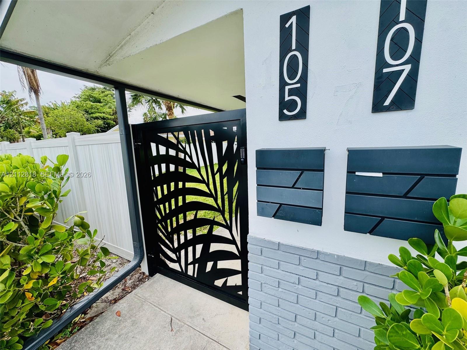 Decorative black metal gate with leaf-pattern cutouts, matte black wall-mounted mailboxes, vertical modern address signage, and a textured grey brick masonry accent wall