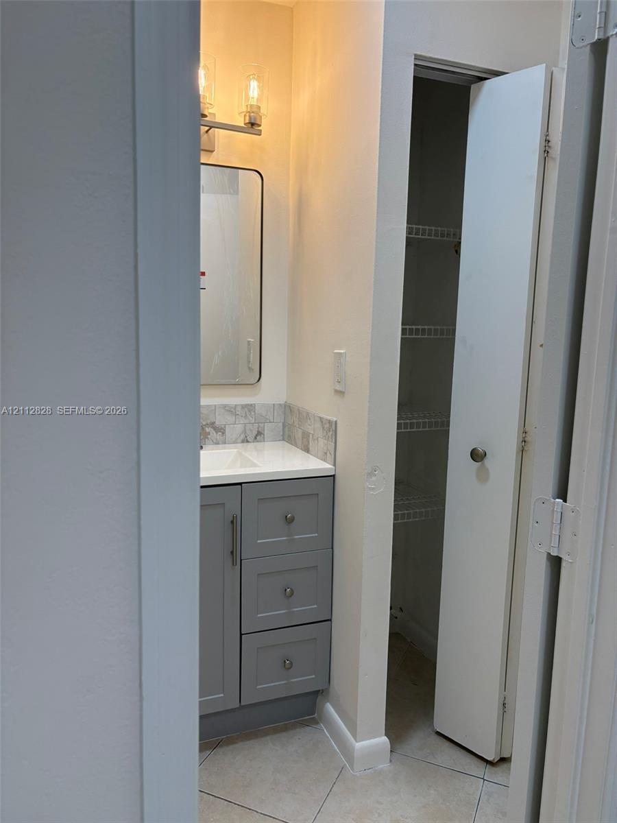 Gray vanity cabinet with a white countertop, tiled backsplash, rectangular mirror with metallic frame, two-light fixture, and built-in closet with wire shelving