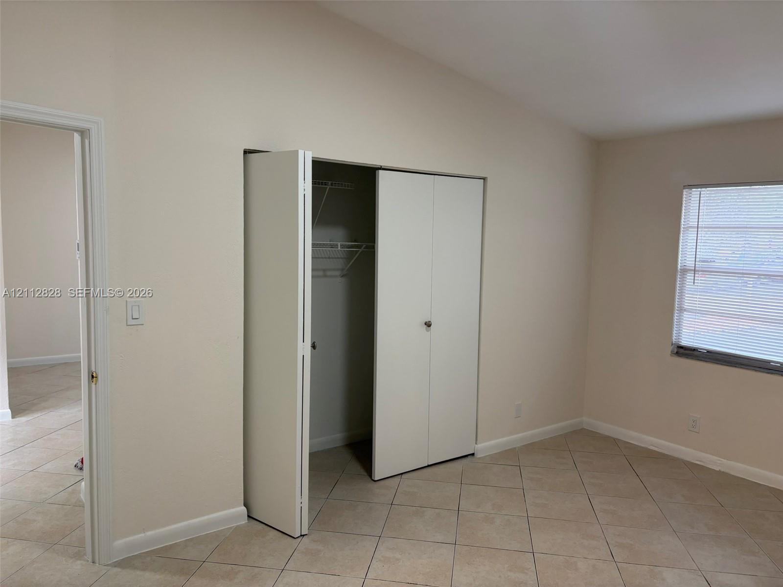 Interior room featuring neutral wall tones, large-format tile flooring, a closet with bifold doors and integrated shelving, and a window with horizontal blinds