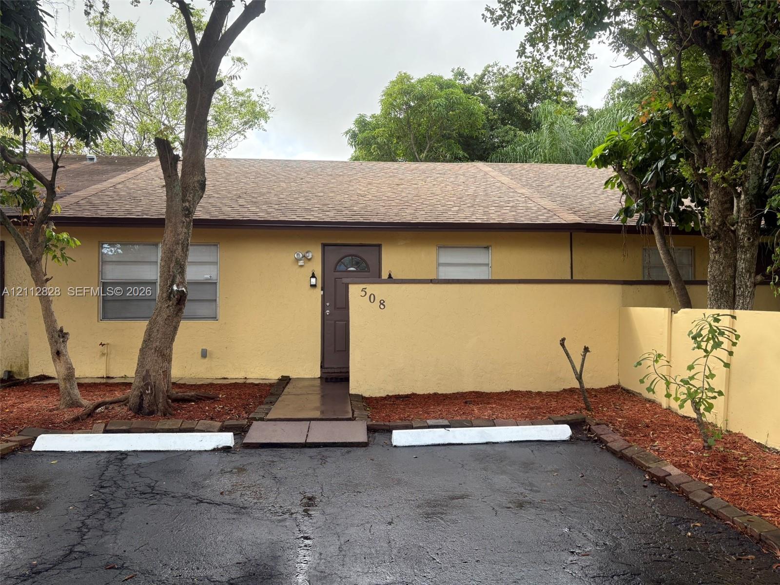 Single-story residence featuring a textured exterior, shingled roof, solid entry door, privacy wall, and paved surface parking