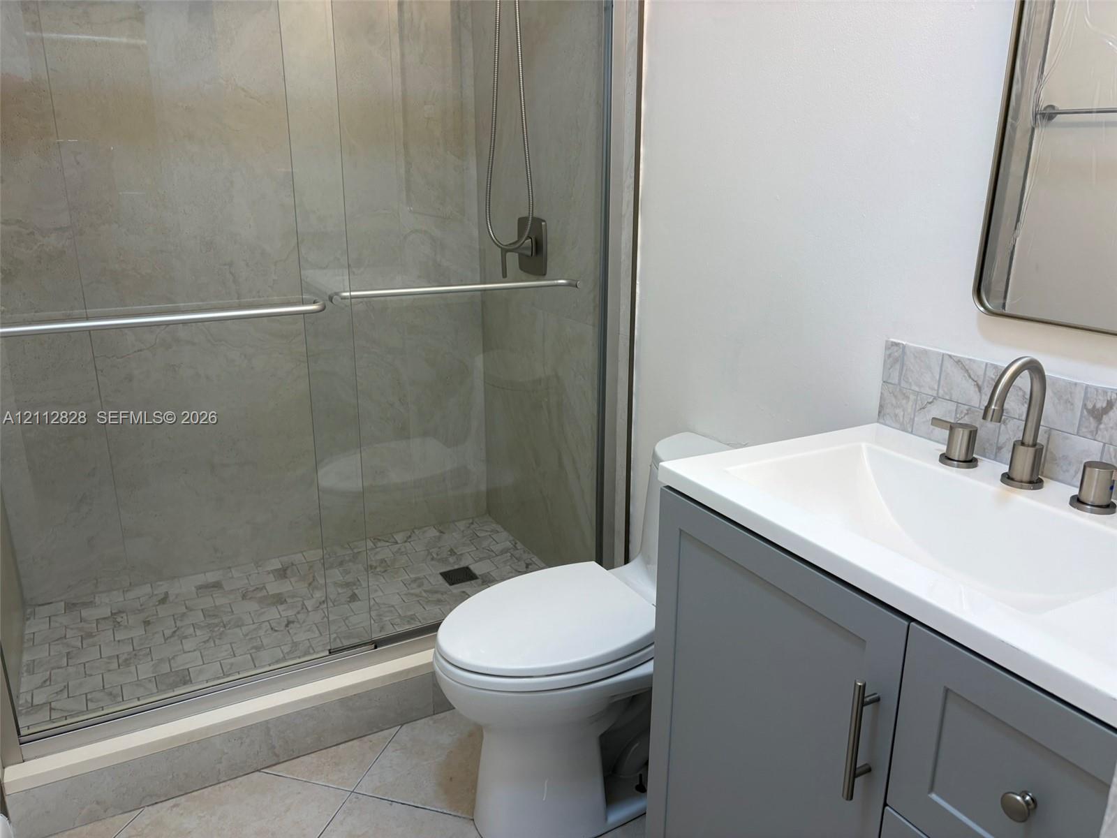 Bathroom featuring a walk-in shower with sliding glass doors, a vanity with a white countertop and undermount sink, stone-finish backsplash, and a wall-mounted mirror