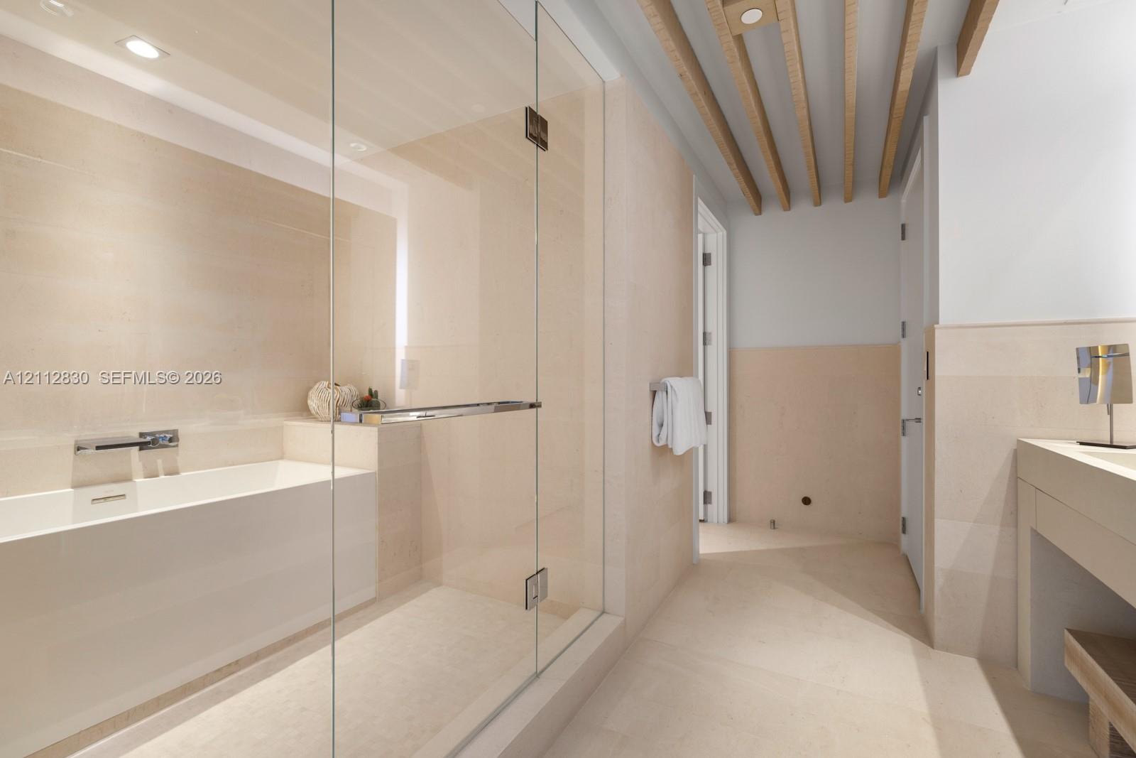 Bathroom featuring a glass-enclosed shower, soaking tub with wall-mounted faucet, neutral stone-finish wall and floor tile, and exposed wooden ceiling beams