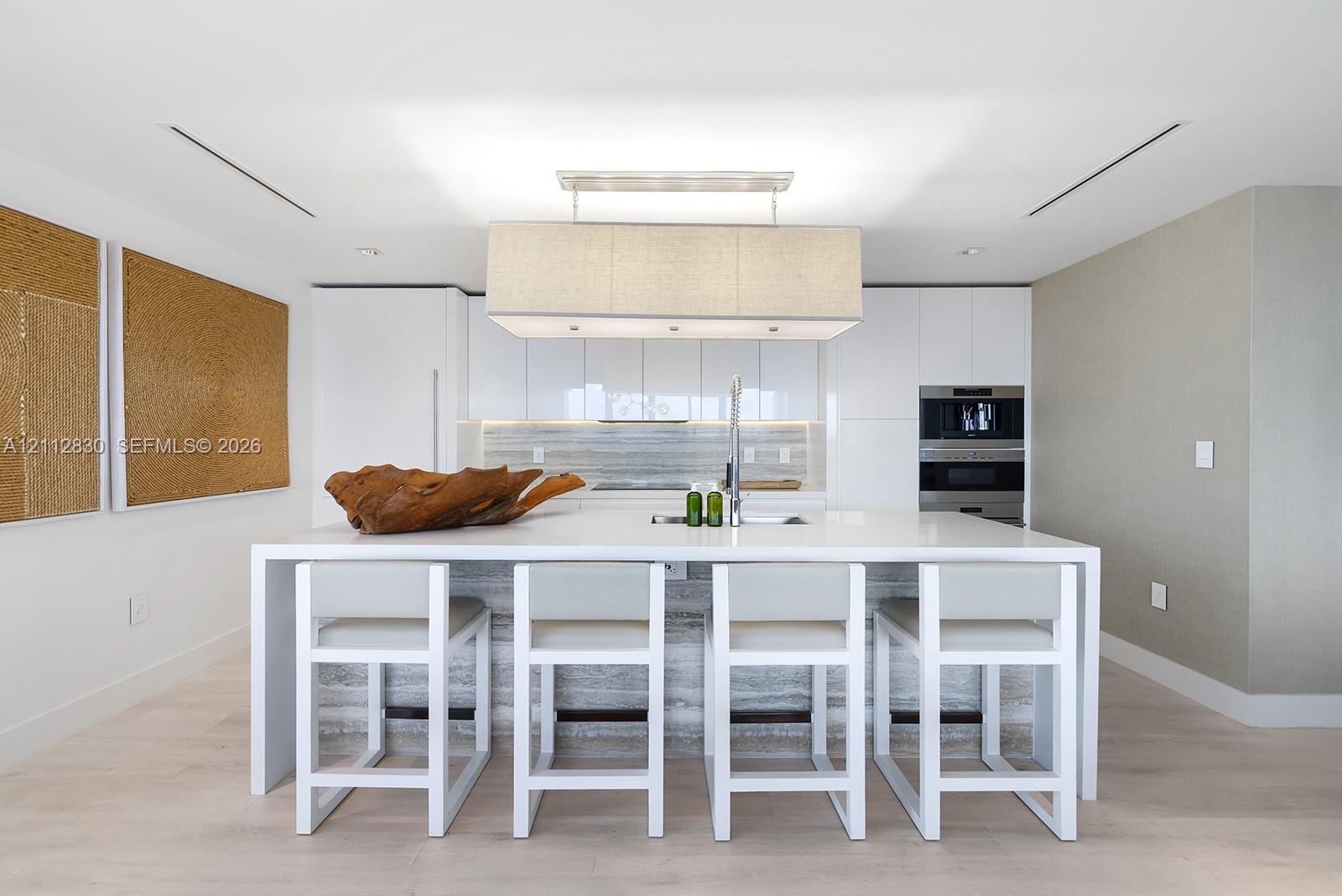 Kitchen featuring high-gloss cabinetry, stone-finish island waterfall countertop, integrated stainless steel appliances, and rectangular pendant light fixture over a breakfast bar