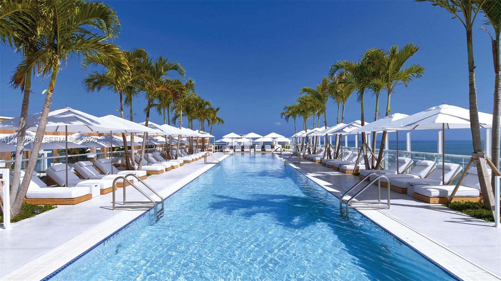 Linear swimming pool featuring white tile decking, stainless steel entry rails, perimeter palm trees, and ocean horizon backdrop