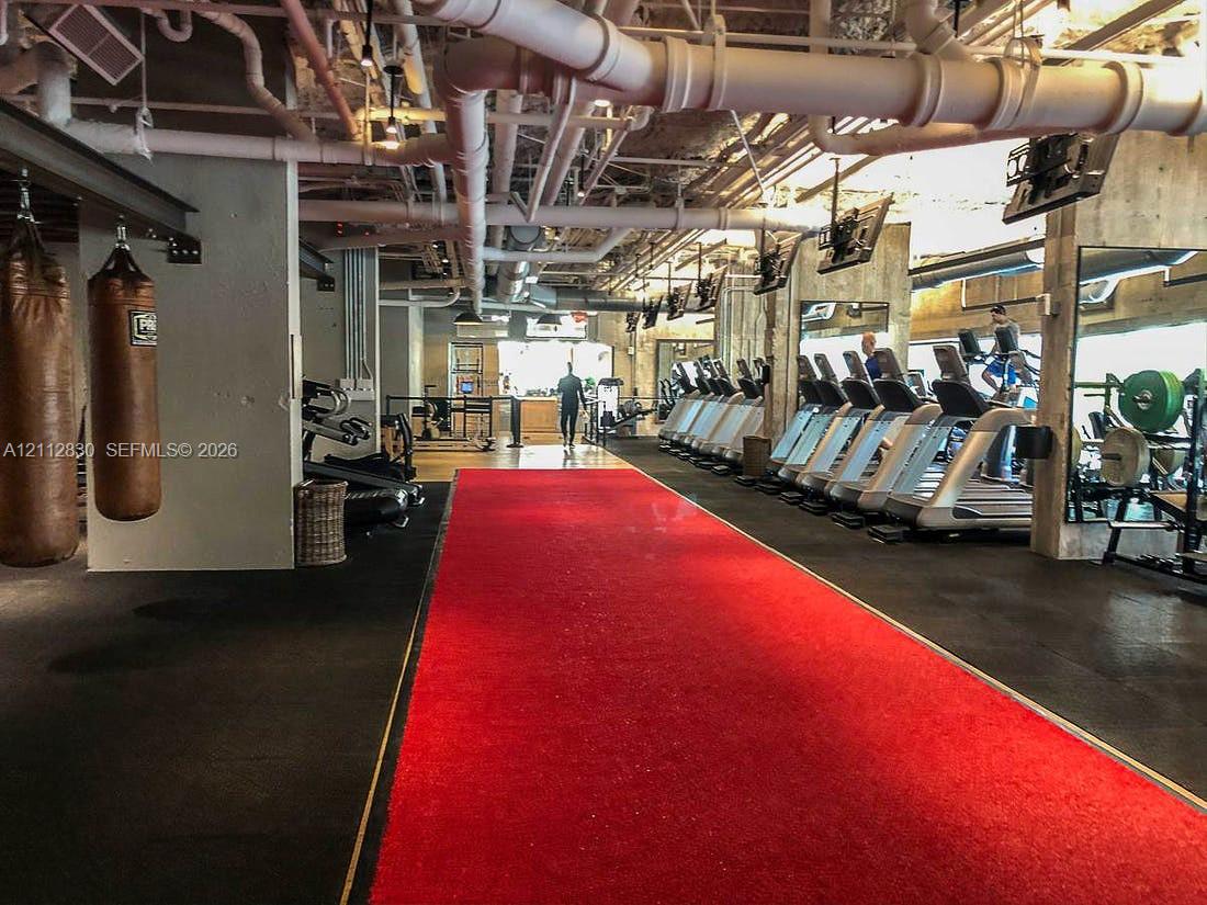 Industrial fitness space featuring exposed ceiling infrastructure, rubberized flooring, a centered synthetic turf track, and full-wall mirrors