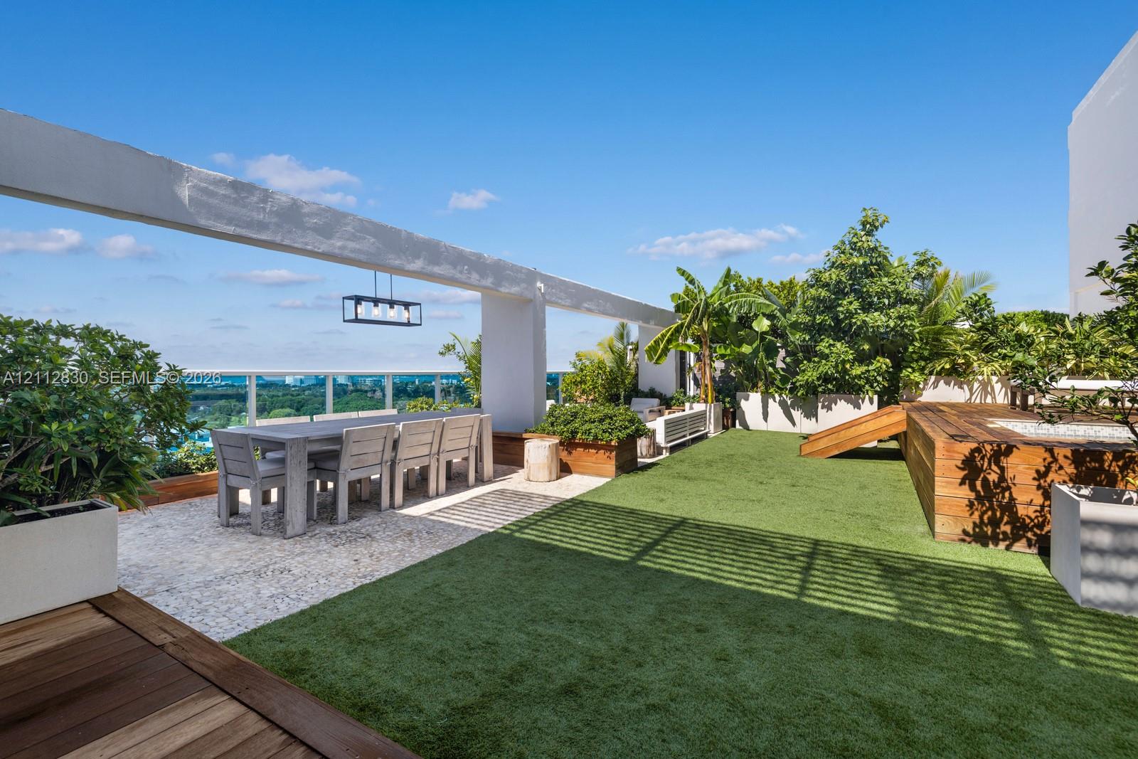 Rooftop terrace featuring synthetic lawn, wood-decking accents, stone-tile flooring, glass panel railing, and overhead structural beam with integrated pendant lighting