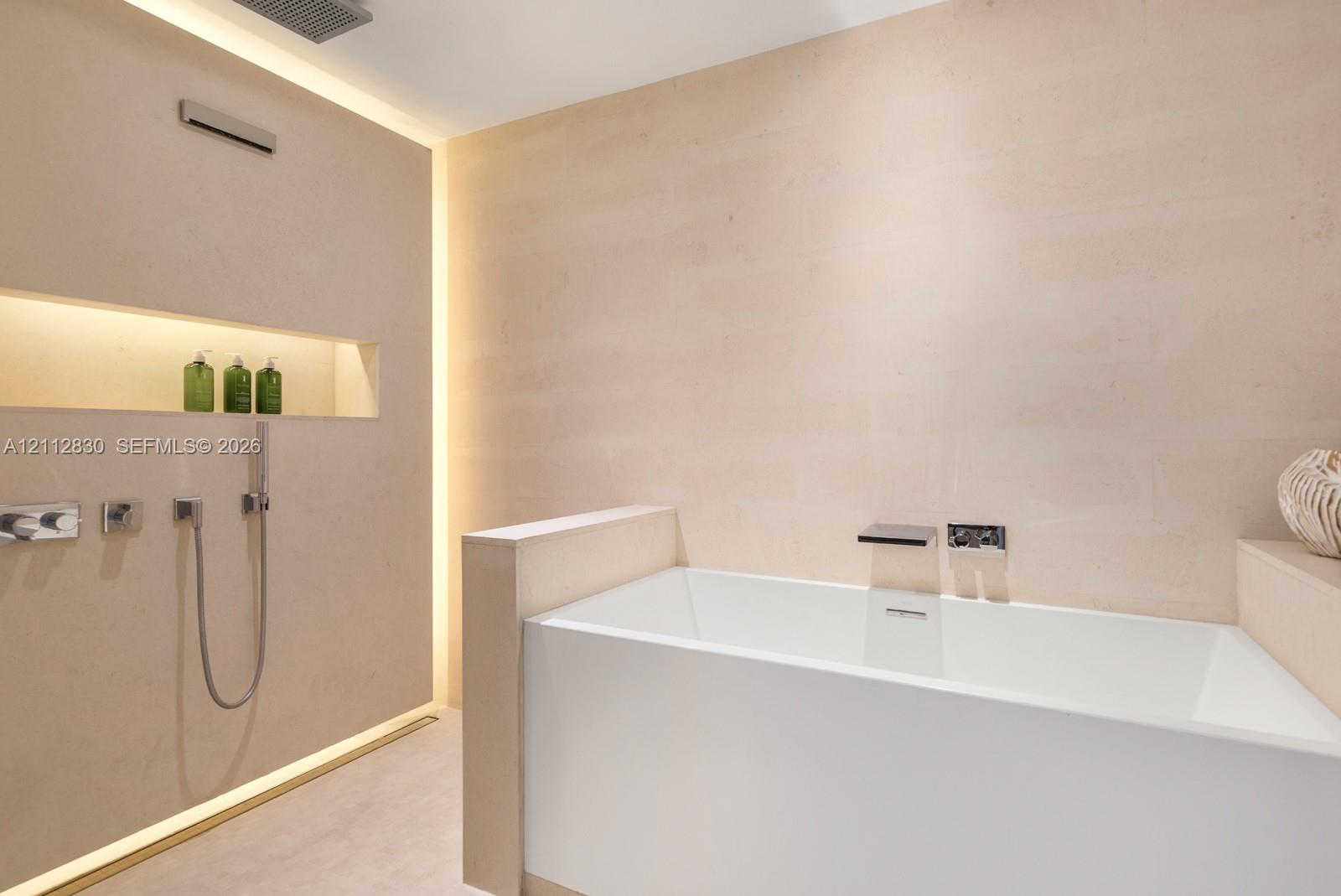 Freestanding deep-soaking bathtub with wall-mounted waterfall filler, integrated stone-finish wall cladding, recessed wall niche, concealed perimeter accent lighting, and polished chrome hand-held shower fixture