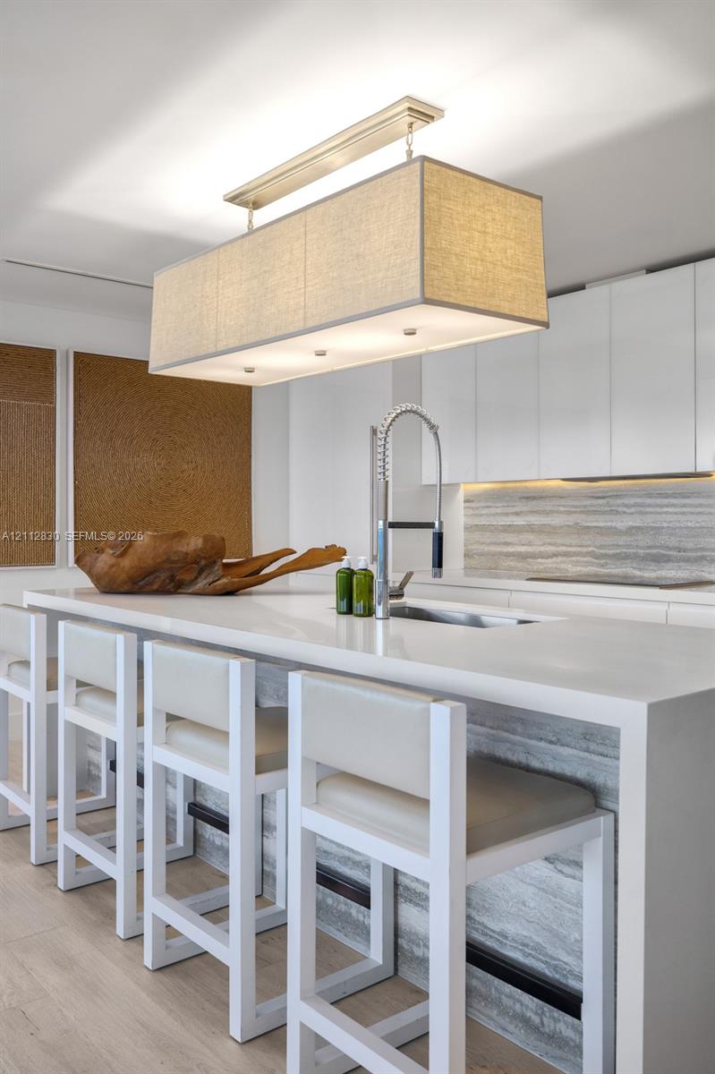 Kitchen island featuring a waterfall-edge countertop, stone-tiled base, undermount sink with a gooseneck faucet, white flat-panel cabinetry, and a rectangular fabric-shade pendant light fixture